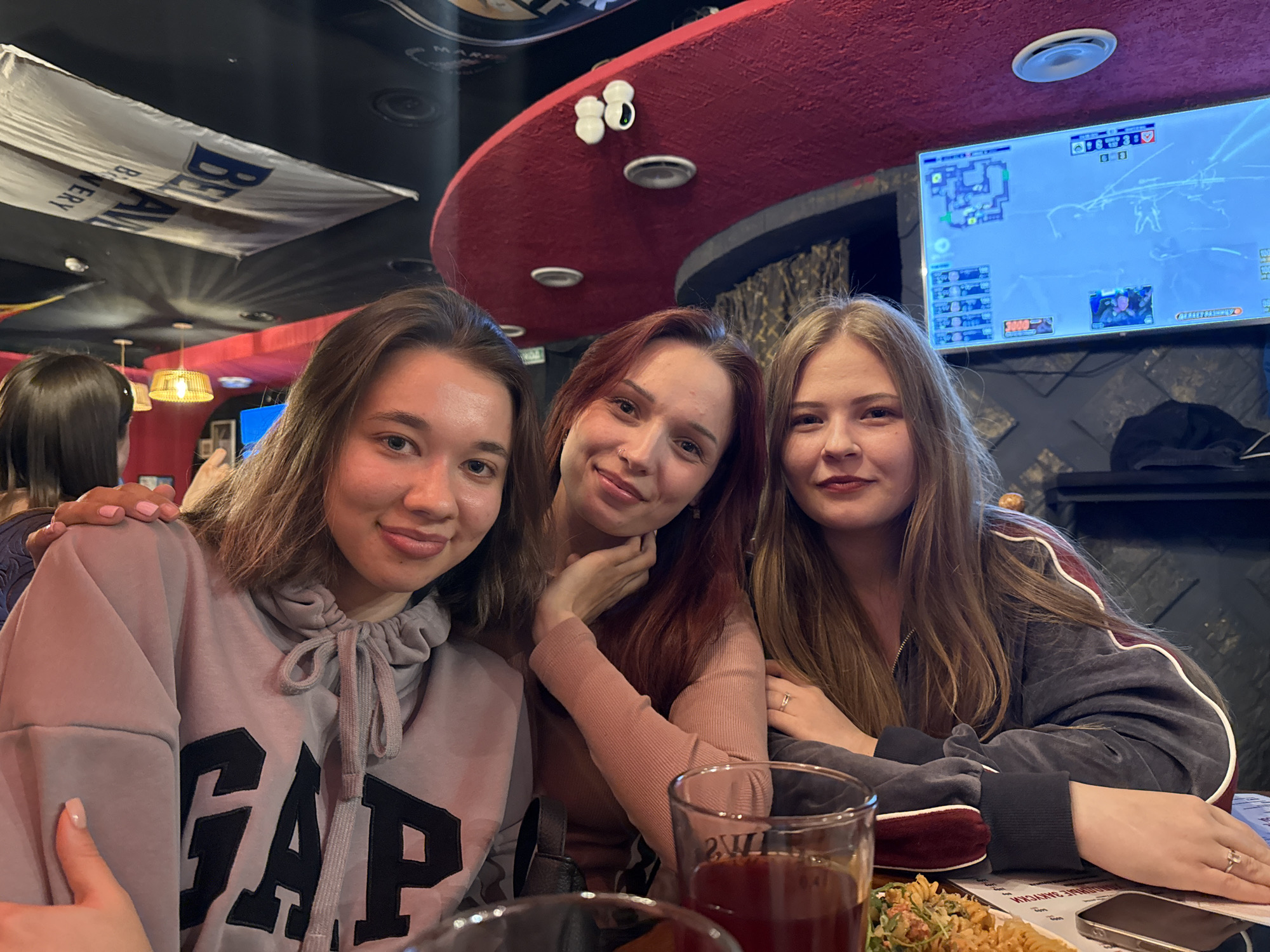 Three young women are sitting together at a table inside a bar or restaurant. The woman on the left wears a grey GAP sweatshirt and has light brown hair. The woman in the middle has reddish-brown hair and wears a pink long-sleeved shirt. The woman on the right has blonde hair and wears a dark grey sweatshirt with a red stripe. They all have fair skin and appear to be smiling or in good spirits. There are glasses of liquid on the table in front of them, along with a plate of food and a mobile phone. Behind them, a large TV screen displays a video game. Overhead, there is a red, curved structural element and a banner is hung up. The banner reads "BELARUSIAN BREWERY". The location is most likely Chaikovsky, Russia, based on information related to the brewery.