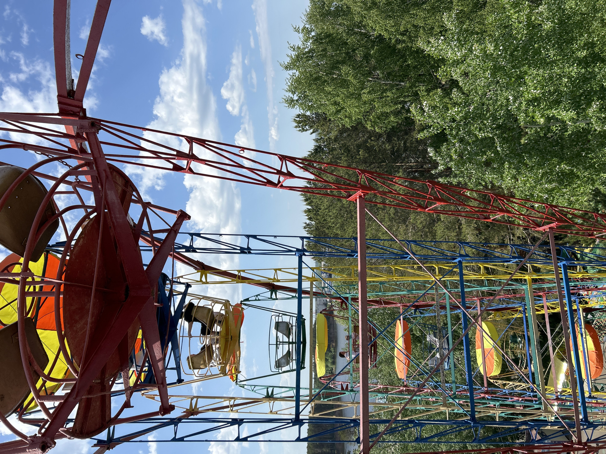 The image depicts a colorful Ferris wheel in the city of Chaikovsky, Russia, as the subject is a Ferris wheel painted in various colors, including red, yellow, blue, and orange. The carriages appear to be in a vintage style, with a few occupied by individuals. The surrounding environment includes trees and a body of water, possibly a lake or river. The sky is partially visible and is blue with scattered clouds. The image is taken from a low angle, emphasizing the height and structure of the Ferris wheel.