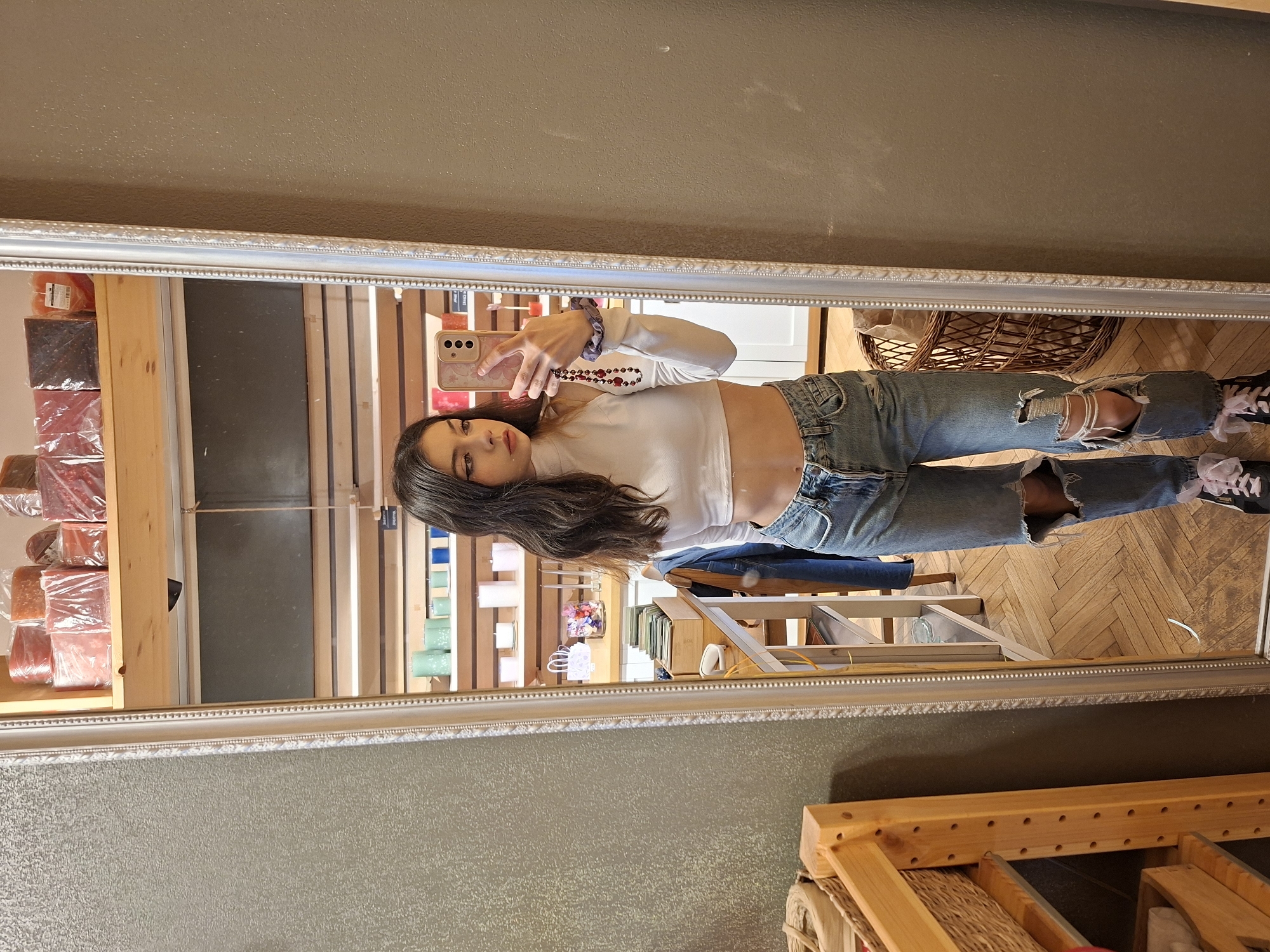 A young woman is taking a selfie in a mirror. She is wearing a long-sleeved white crop top, ripped blue jeans, and sneakers with pink laces. Her dark hair is long and loose. She is holding a cell phone in her right hand. The mirror is in a room with wooden floors and shelves of various items. A ladder and baskets are also visible in the background. The store is likely located in Sofia, Bulgaria.