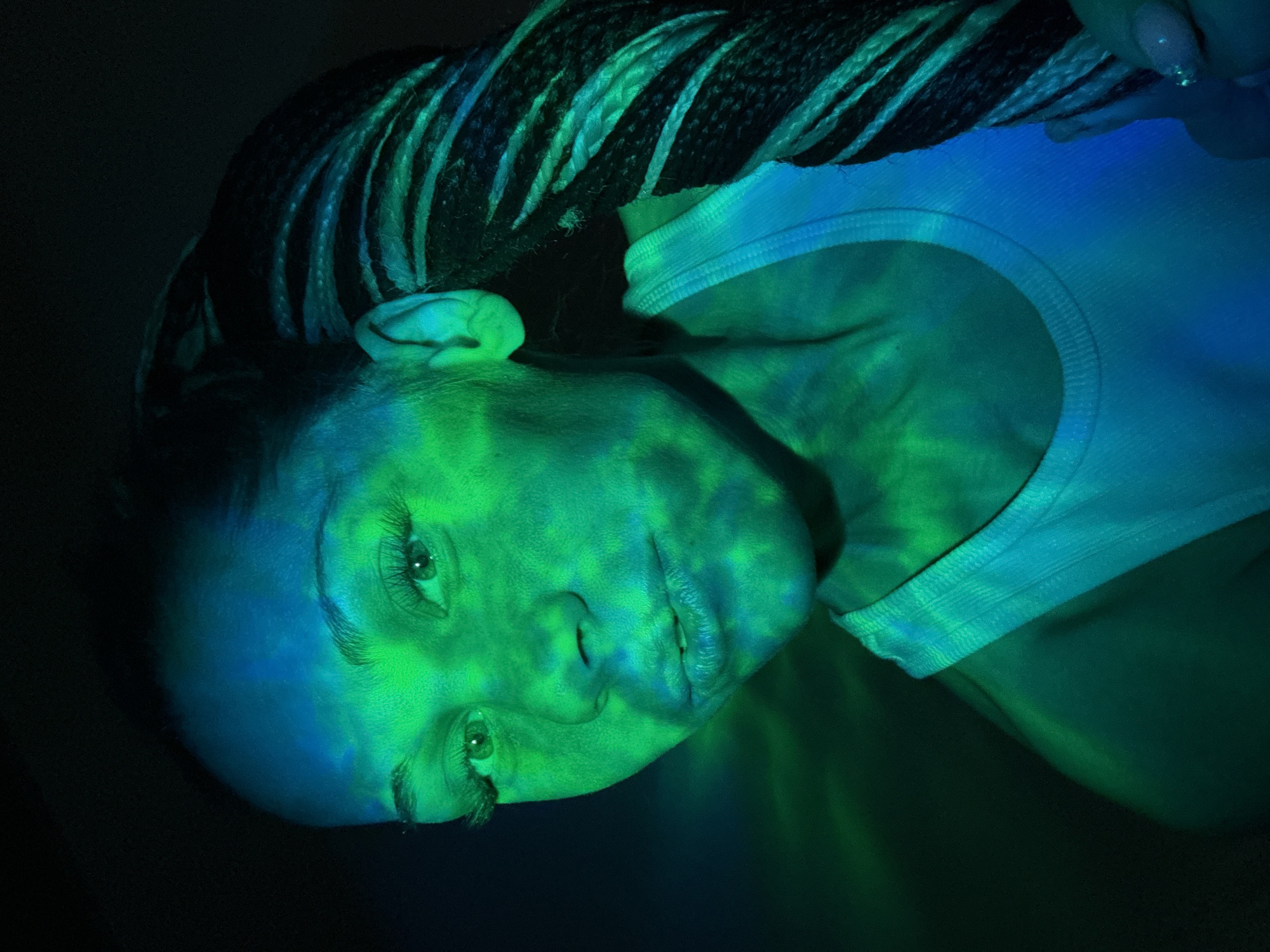 The media file is a portrait of a person, likely female, captured in a dramatically lit, low-light setting. The subject's head is tilted, and she appears to be lying down or leaning back, looking towards the camera. Her face, neck, and upper chest are illuminated by vibrant, shifting patterns of green and blue light, resembling reflections from water or a projector, creating an abstract and ethereal effect. She has dark dreadlocks or braided hair, which also show streaks of blue and green. A light-colored top, possibly a tank top, is visible on her right shoulder and chest. In the upper right corner, a hand with manicured nails is partially visible, seemingly interacting with her hair or an unseen element. The background is dark and indistinct, focusing attention on the illuminated subject. Her expression is calm and contemplative, with her eyes open. The artificial lighting suggests an artistic photoshoot, a club, or a performance event. There is no visible text in the image. The scene is set in Rusanovo, Russia.