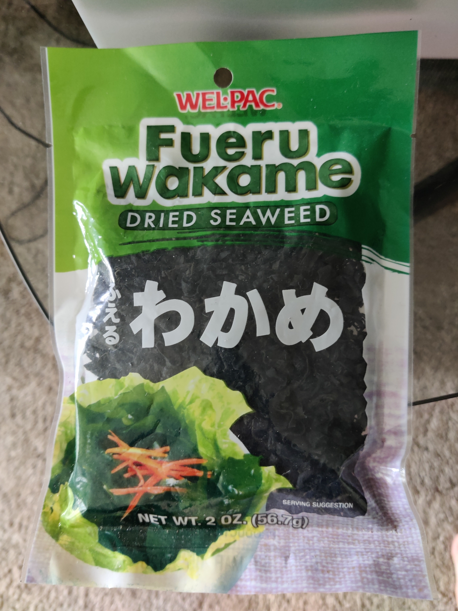 A product image displays a green plastic pouch of Wel-Pac Furu Wakame Dried Seaweed. The pouch is oriented vertically, with the top facing upwards. The branding "WEL-PAC" is in red capital letters, followed by "Fueru Wakame" in large green bubble letters with white outlines. Below this, "DRIED SEAWEED" is written in green text on a white background. The majority of the pouch is filled with dark, dried seaweed. Towards the bottom, an image shows a serving suggestion with green lettuce leaves and julienned orange carrots. Text at the bottom of the pouch indicates "SERVING SUGGESTION" and "NET WT. 2 OZ. (56.7g)". The background appears to be a textured surface, possibly a carpet or rug, in a neutral color. The image is a close-up of the product, with no discernible environmental context within Lower Paxton Township, United States of America.