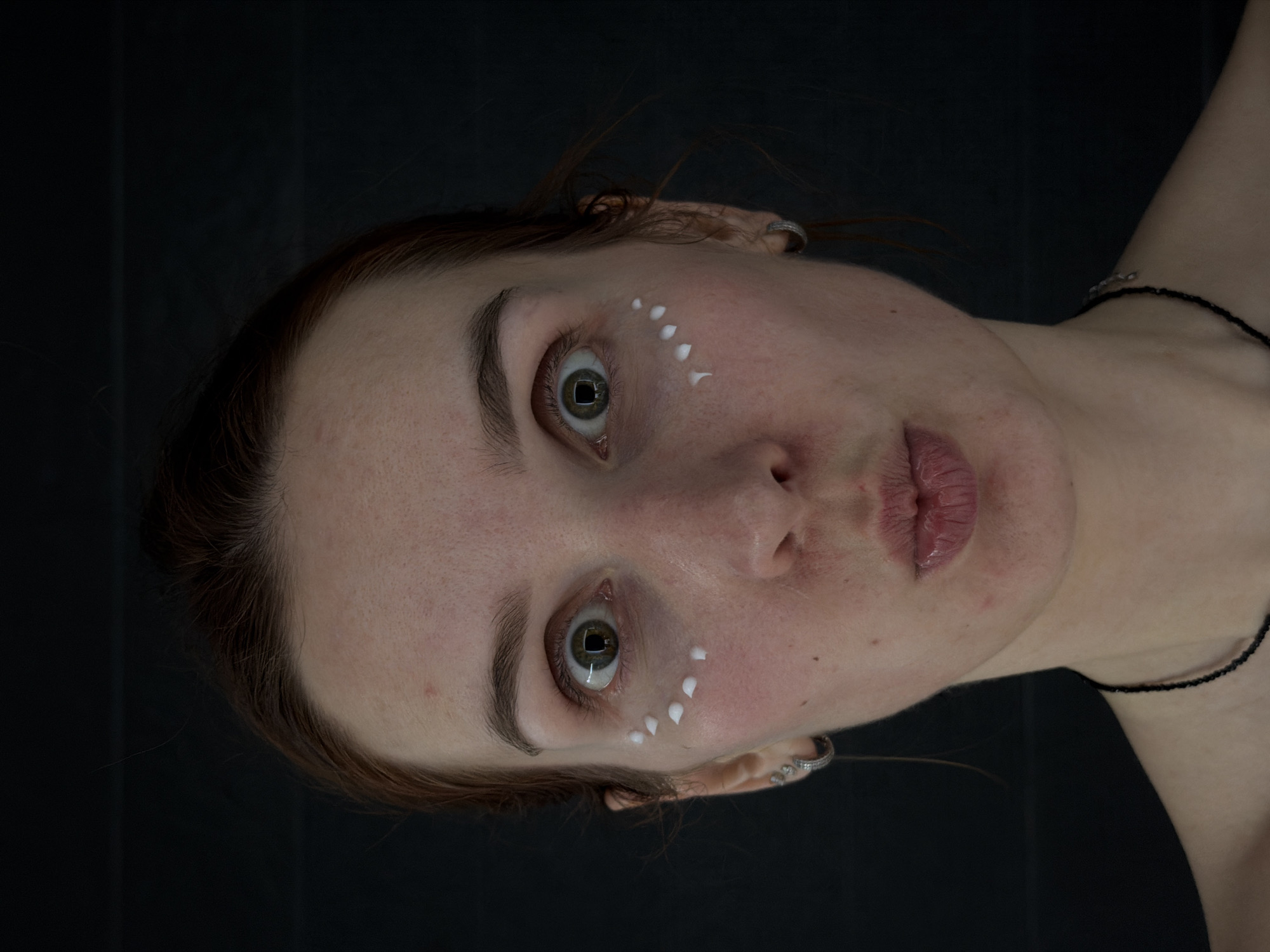 A close-up portrait shows a fair-skinned woman with reddish-brown hair, light green eyes, and a necklace. White cosmetic dots are applied near her eye, in front of her ear. She appears to be facing up at the camera at an angle. The background is dark. There is no indication the image was taken in Chaikovsky, Russia.