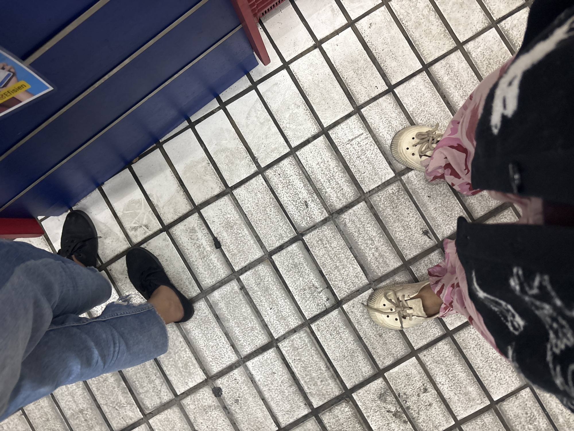 The image shows a low-angle view of two individuals standing on a tiled floor. The person in the foreground is wearing off-white canvas sneakers and brightly colored pink and black patterned pants. The person in the background is wearing blue jeans and black slip-on shoes. The tiled floor is light-colored with dark grout lines. A dark blue structure with some yellow signage is visible to the right of the frame, and a red chair leg is partially visible. The scene appears to be an indoor public space. There is no visible indication of the specific location in North Jakarta, Indonesia.