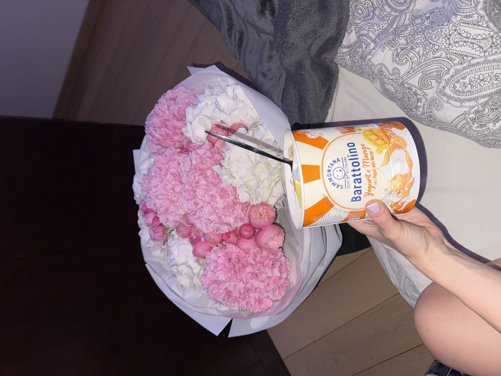 A close-up shot shows a bouquet of light pink and white flowers, wrapped in white paper, placed next to a container of "Barattolino" yogurt with a mango flavor. A silver spoon rests in the center of the bouquet. A hand holds the container of yogurt. The scene is set on a bed with a gray patterned pillow. Parts of the dark brown floor are visible around the bed. There is no indication that the scene is located in Warsaw, Poland.