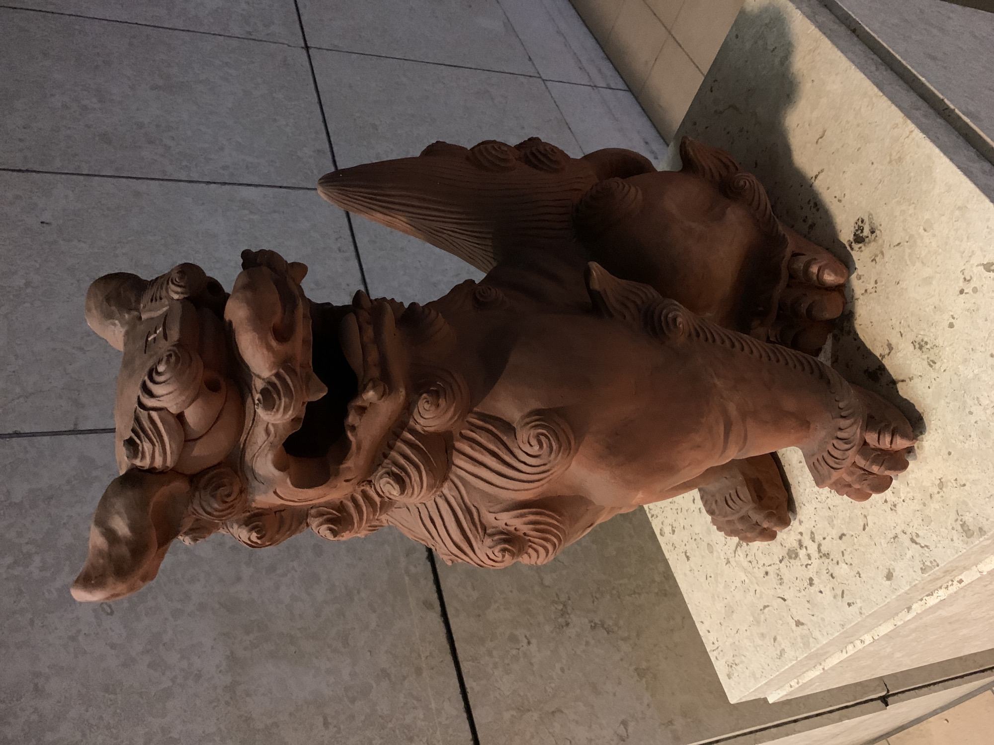 This media file depicts a clay sculpture of a mythical creature, likely a Komainu, a Japanese lion-dog statue. The sculpture is reddish-brown in color and appears to be in motion, with its mouth open and its body in a leaping or charging pose. The creature has a mane composed of intricately carved swirls and lines, and its paws are detailed with individual toes. It is positioned on a light-colored, textured surface that resembles a stone pedestal or platform, which is set against a tiled floor. The lighting suggests an indoor setting with artificial light casting shadows, particularly a prominent shadow on the pedestal to the right of the sculpture. The overall impression is one of a guardian or decorative piece, common in Japanese architecture and garden design. The specific location cues of Naha, Japan, suggest this sculpture could be found at a temple, shrine, or public space within the city. There is no visible text in the image.
