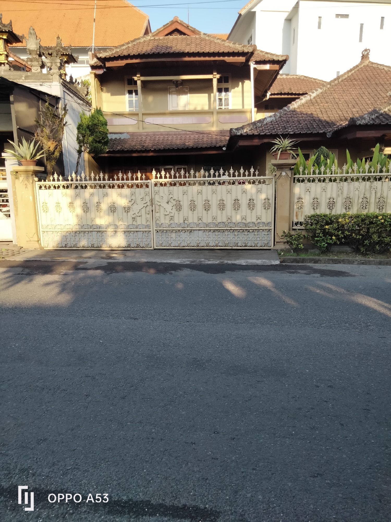 The image shows a residential street in Denpasar, Indonesia, with a focus on the gate and facade of a house. The house has a light yellow exterior and a traditional tiled roof. The gate is made of ornate white metal with decorative patterns and lion motifs. In front of the gate, there is a paved sidewalk and a road. The street is lined with vegetation, including bushes and potted plants. The lighting suggests it is either early morning or late afternoon, with long shadows cast across the scene. The sky is clear and blue. The OPPO A53 watermark is visible in the lower left corner of the image.