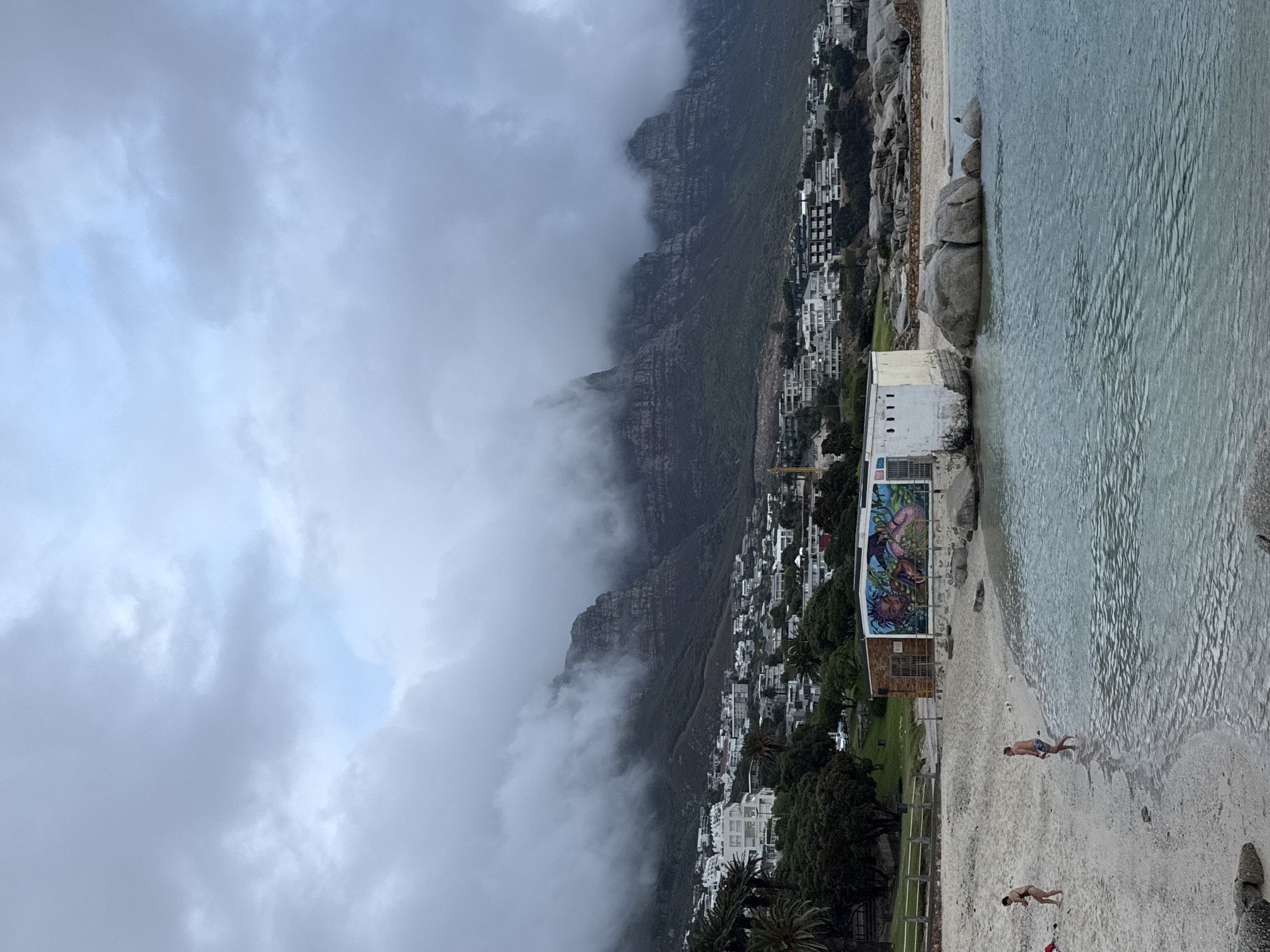 The image captures a beach scene near Cape Town, South Africa. In the foreground, a sandy beach meets the ocean, with two individuals wading in the water. Adjacent to the beach, a structure with a mural is visible. Behind the beach and structure, a mountain range covered in fog rises, with buildings and homes dotting the lower slopes. The sky is overcast.