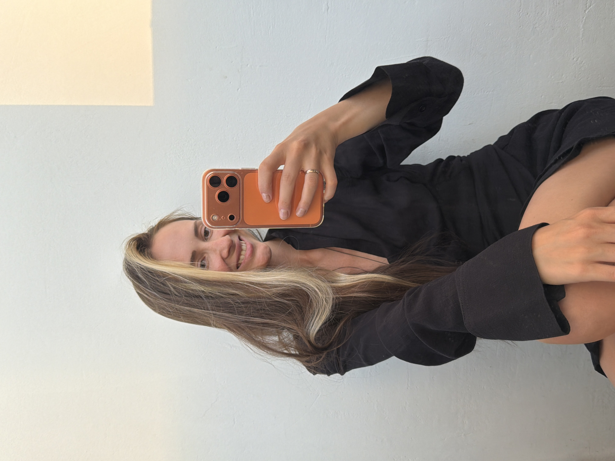 A woman with long brown hair with blonde highlights takes a selfie. She's wearing a black top, possibly a dress or shirt, and holding an orange smartphone. The background is a white wall with a patch of lighter color in the upper left corner. There is no visible evidence to confirm this image was taken in Khimki, Russia.