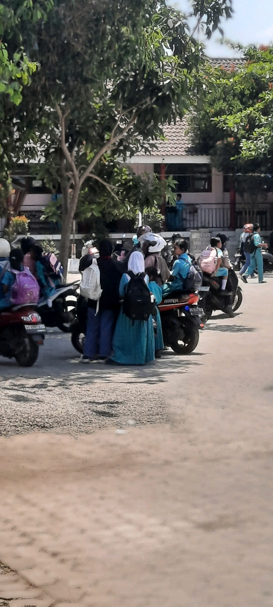 In Tangkil, Indonesia, a group of students wearing school uniforms and hijabs are gathered around parked motorcycles. Some students are wearing backpacks. The scene appears to be outside a school building, with trees and a tiled roof visible in the background. The ground is paved with asphalt, and there are shadows cast by the trees. The overall atmosphere suggests a typical school day or dismissal.