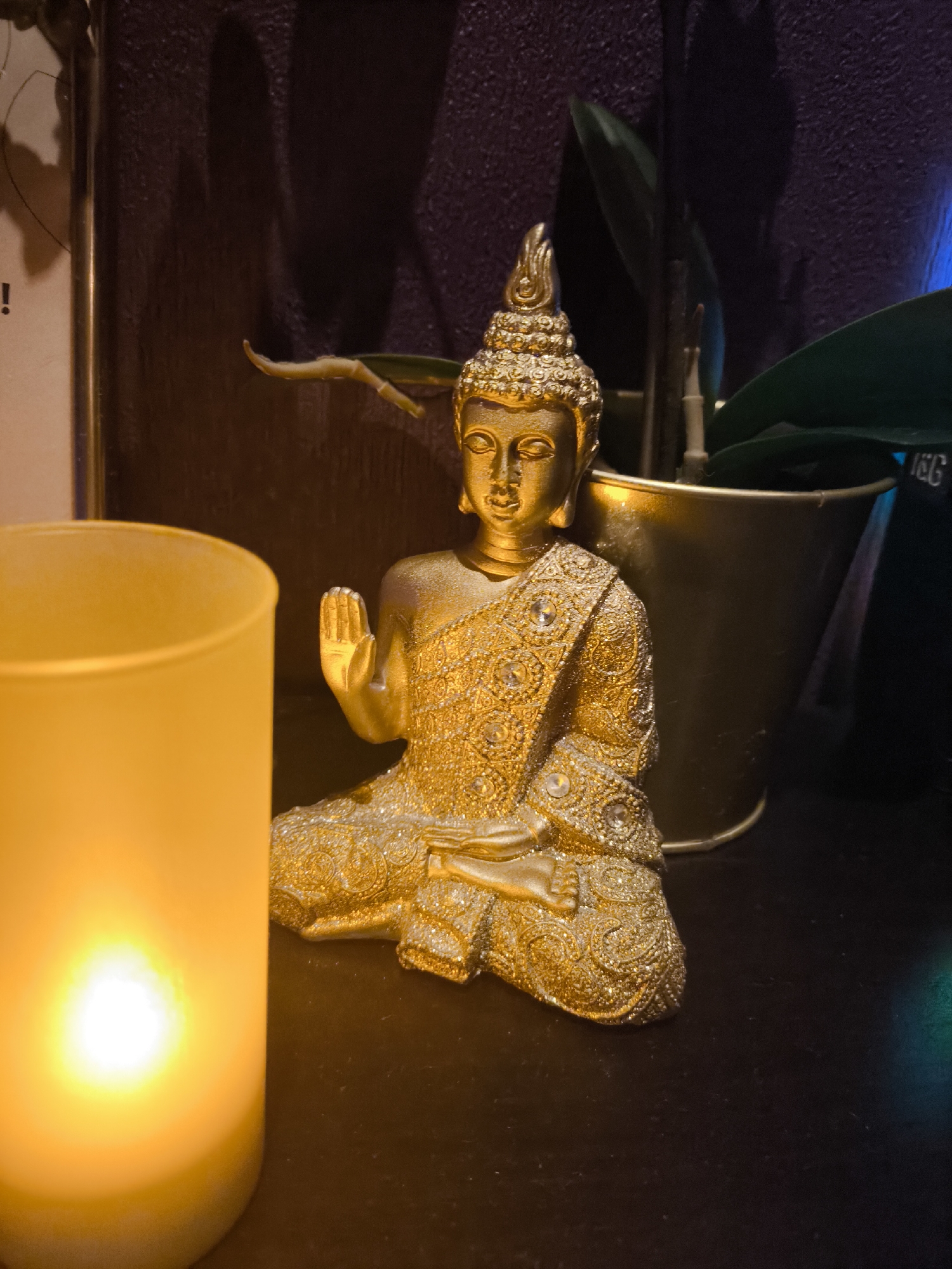 A close-up shot of a golden Buddha statue in a meditative pose with a raised hand, placed next to a lit yellow candle. Behind the statue, there's a gold-colored plant pot with green leaves. The background is dark, with a textured purple wall visible. There's no indication of this being in Khimki, Russia. The objects are arranged on a dark surface.