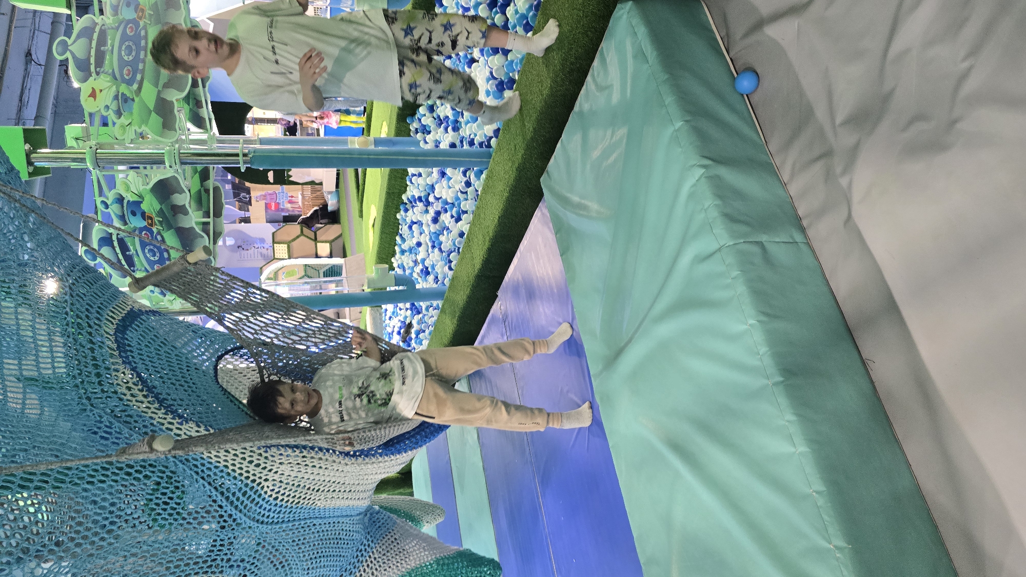 This is a photograph of an indoor children's play facility, likely located in Izhevsk, Russia. The scene features two male children engaged in recreational activities. On the left, a younger boy, approximately 6-8 years old, with short dark hair, stands within a grey net hammock suspended from a large, intricately woven blue, white, and teal net climbing structure. He wears a light-colored t-shirt with a graphic design (partially visible text "SEAL AND...") and light brown sweatpants with dark zippers near the pockets, along with white socks. He looks towards the camera with a slight smile, his hands lightly gripping the net. To the right, an older boy, approximately 10-12 years old, with short light brown hair, is in mid-stride, walking on a raised, narrow strip of artificial green grass. He wears a white short-sleeved t-shirt with dark text and patterned shorts (white with dark blue star motifs), along with white socks. His left foot is elevated, indicating motion. This green strip borders a large ball pit filled with numerous small plastic balls in shades of white, light blue, and dark blue. The immediate foreground features various padded floor mats: a light blue rectangular mat, a darker blue padded section, and a prominent light teal-colored padded mat in the lower right, which abuts a grey floor. A small blue plastic ball rests on this grey floor next to the teal mat. In the background, beyond the ball pit, additional playground structures are visible, including vertical light blue poles and large, decorative green and blue cutouts resembling crowns or castles, suggesting an elaborate play environment. Areas of green turf, possibly for miniature golf, are also discernible in the deeper background. The facility is brightly lit, consistent with an indoor recreational space.
