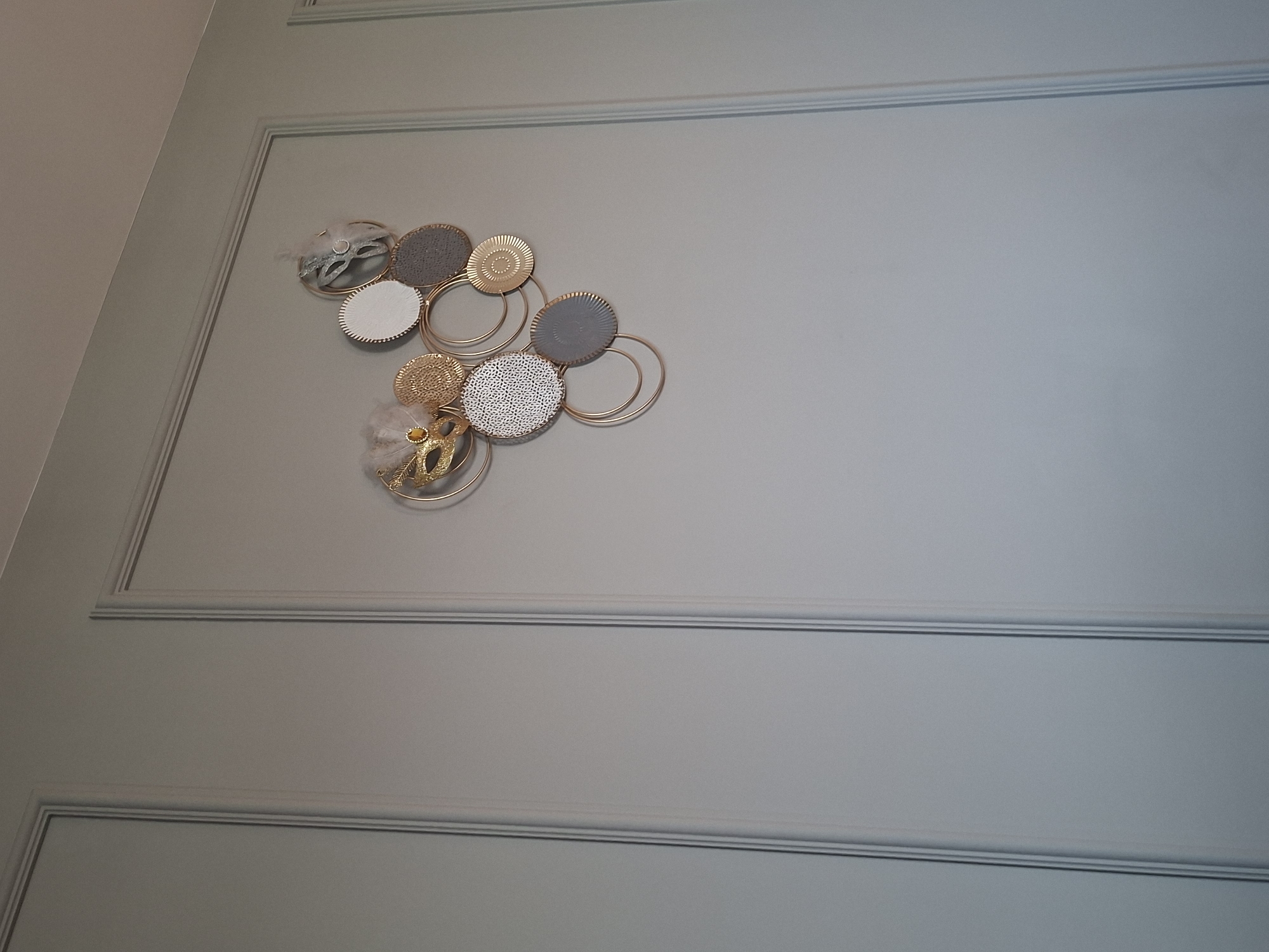 The image shows a wall decoration featuring multiple circular elements of varying textures and colors, predominantly gold, white, and gray. Some circles have intricate patterns, while others are solid. The decoration is arranged in a cluster, with some elements overlapping and connected by gold rings. A golden mask with a yellow gem and light-colored feathers is also part of the arrangement. The wall itself is painted a light gray-blue and features vertical paneling with subtle trim. The lighting suggests an indoor environment. The image does not provide any information about the city of Branipole, Bulgaria, or any people or actions taking place.