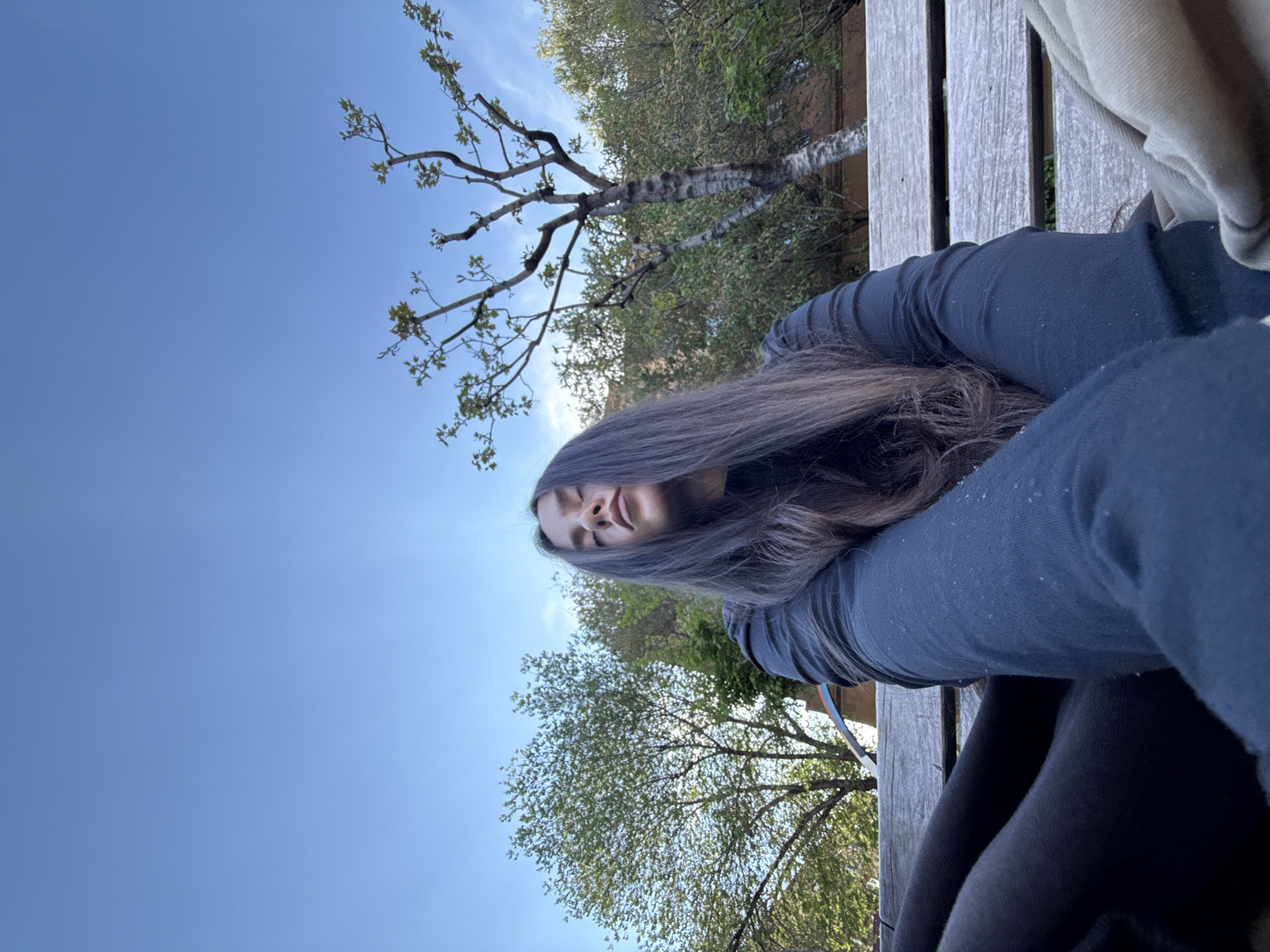A young woman with long, dark hair is lying on a wooden bench outdoors. She is wearing a dark-colored long-sleeved shirt and dark pants. Her head is tilted slightly, and she appears relaxed. The background features a clear blue sky and trees with green foliage. A birch tree is visible. The location is Yerevan, Armenia.