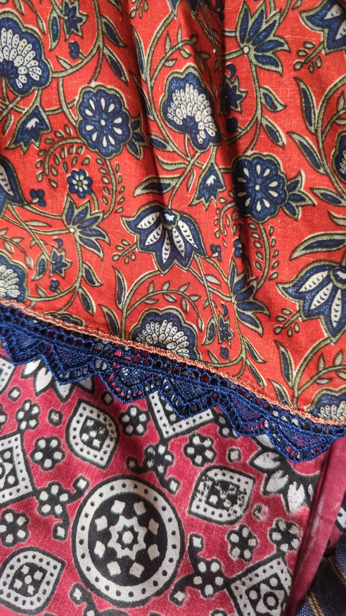 The image displays a close-up view of two sections of fabric, separated by a navy blue lace trim. The upper section of fabric features a red base adorned with a floral pattern in blue and white, with green vine-like elements connecting the flowers. The lower section of fabric has a red base with a repeating pattern of black and white geometric shapes and stylized flowers. The lighting appears to be natural, and the fabric is slightly wrinkled, suggesting it might be part of clothing or home decor. There are no people or specific environmental elements visible that would indicate a location within Wah Model Town phase 1, Pakistan.