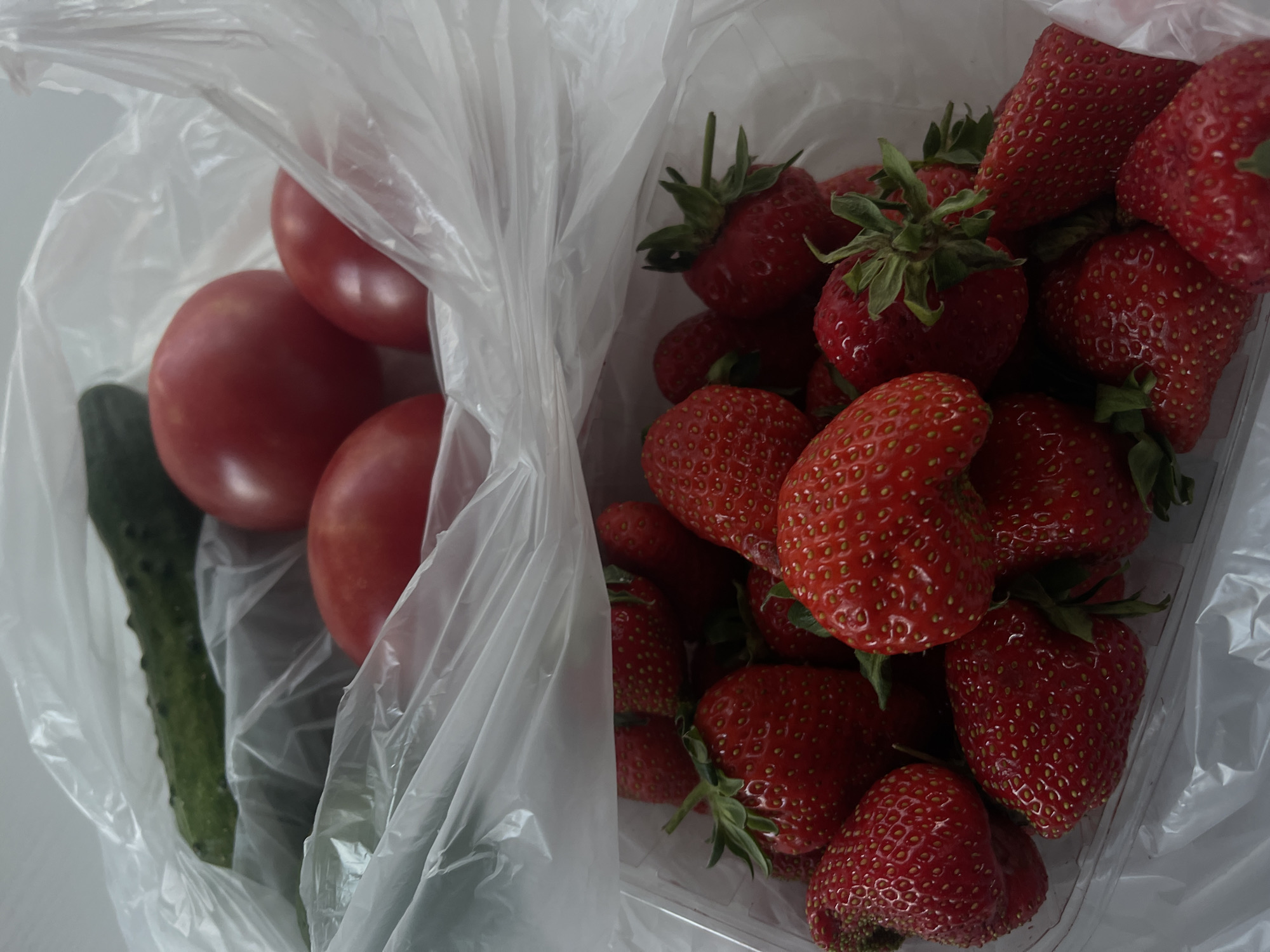 The image depicts a plastic bag containing a cucumber and three tomatoes, placed next to a plastic container full of strawberries. The scene is presumably located in Мосметаллоконструкция Онт, Russia.