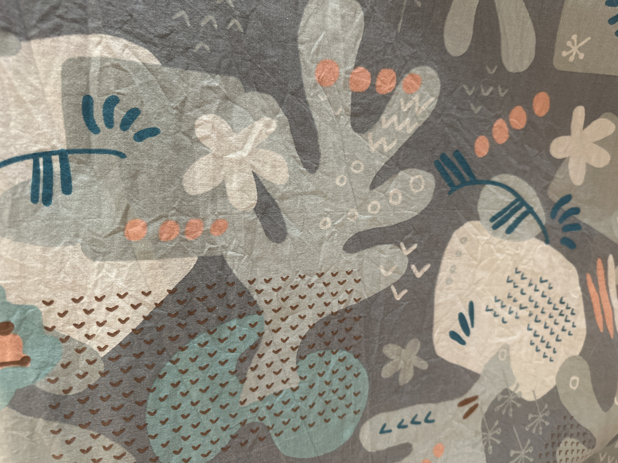 The image shows a close-up, slightly wrinkled fabric with a playful, abstract pattern. The pattern features a variety of organic shapes in muted tones of gray, light beige, and teal, interspersed with small, coral-colored dots and clusters of brown "v" shapes. There are also stylized floral motifs and simple line drawings of what appear to be creatures or abstract figures. The overall impression is one of a child-like, whimsical design. No people, specific objects, or discernible scene are present; the image focuses solely on the textile print. There is no indication of time of day, weather, or a specific location beyond the provided context of Minsk, Belarus, which would be external information. No text is visible in the image.