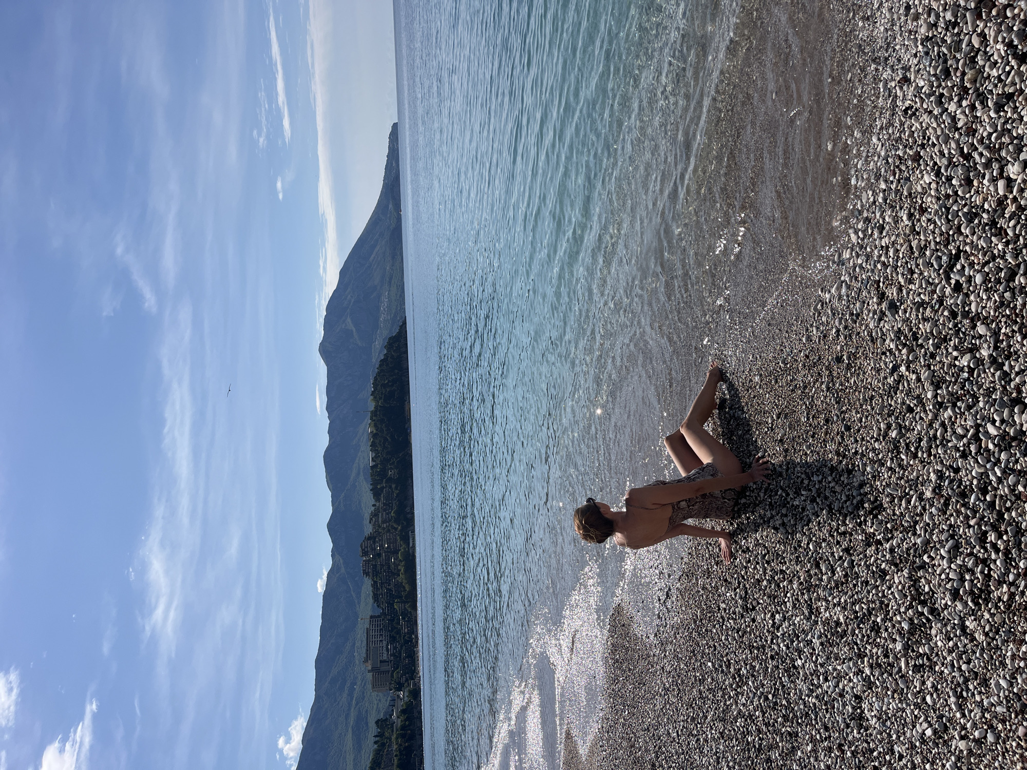 The image presents a serene coastal landscape, likely captured in Budva, Montenegro. In the foreground, a person, appearing to be a woman, is seated or reclining on a pebble beach at the very edge of the water. She is seen from behind, wearing a patterned swimsuit, with her legs extending slightly into the clear, rippling blue water. The sunlight glints brightly off the water's surface, creating sparkling reflections near the shore.
The beach itself is composed of numerous small, smooth, grayish-white pebbles. Beyond the immediate shoreline, a vast expanse of calm, clear blue water stretches out, meeting a dramatic mountainous backdrop. These mountains are rugged, covered in lush green vegetation, and feature several multi-story buildings nestled along their lower slopes, indicating coastal development.
The sky is a clear, vibrant blue with scattered, wispy white clouds. A tiny, distant object, possibly a bird or a glider, is visible high in the sky. The bright and direct lighting suggests it is daytime, likely late morning or early afternoon, with strong sunlight illuminating the entire scene. The overall impression is one of peaceful natural beauty, combining the clear waters and distinctive pebble beach with the imposing mountain scenery characteristic of the Montenegrin coast. There is no visible text in the image.