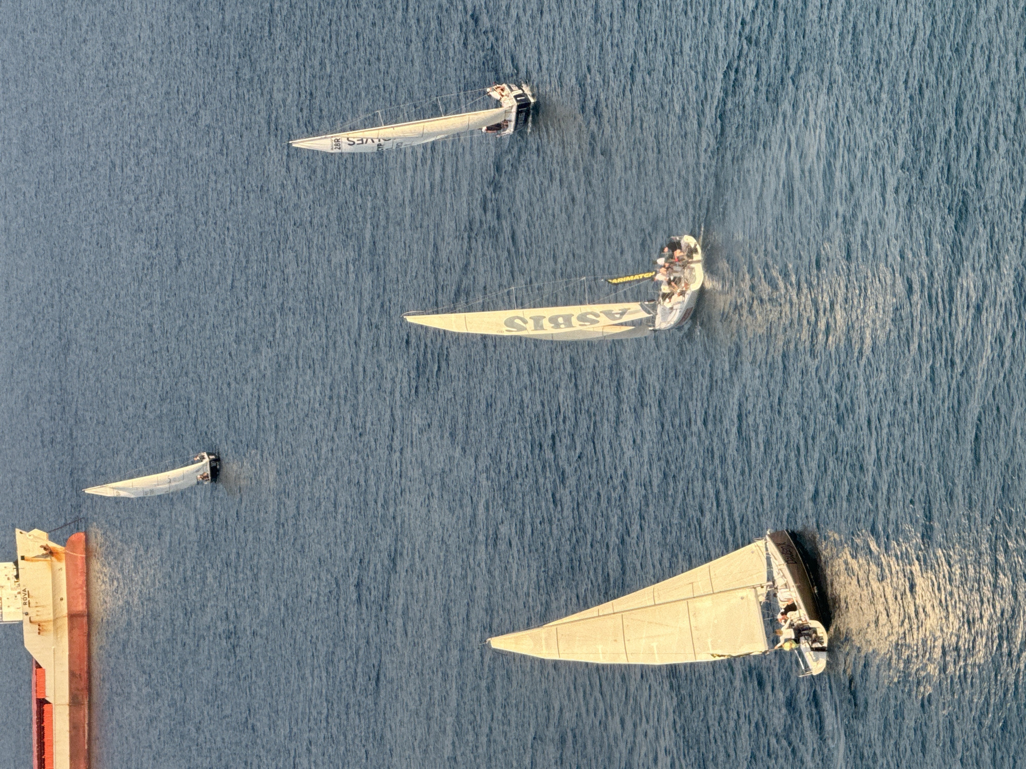 The image shows a wide shot of a body of water, likely the sea, with several sailboats and a large cargo ship. The water is a deep blue, with small ripples on the surface. The cargo ship is in the upper left corner of the image and appears to be docked or anchored. It is a large vessel with a white hull and a red section along its side. There are four sailboats visible. One is in the upper middle of the frame, sailing away from the camera. Another is in the middle right, also sailing away. A third sailboat is in the lower left, sailing towards the camera. This sailboat has a yellow sail with the word "ASBIS" written on it in large letters. The fourth sailboat is in the middle left, sailing towards the camera. The sails of the sailboats are white and appear to be made of fabric. There are people visible on the decks of the sailboats, but their features are not discernible. The lighting suggests it is daytime, with the sun casting reflections on the water. This scene is consistent with the coastline of Limassol, Cyprus, known for its busy port and sailing activities.