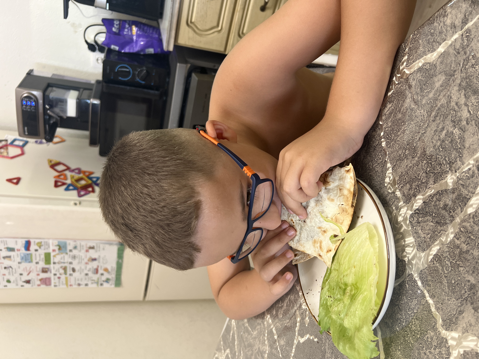 This media file captures a close-up, top-down perspective of a young child eating in what appears to be a kitchen or dining area. The image is rotated 90 degrees clockwise from its natural orientation.
**Subjects:** The primary subject is a child with short, light brown hair, wearing blue-framed glasses with orange accents. Their face is partially obscured as they lean over a plate to eat. Their bare arms are visible, resting on the table.
**Objects:** The child is holding a piece of flatbread, possibly lavash, in both hands, bringing it towards their mouth. On a white plate with a brown rim, there's another piece of flatbread and a large leaf of fresh green lettuce. The plate rests on a dark, glossy table with a distinct white, marble-like or cracked pattern. In the background, various kitchen elements are visible: light-colored wooden cabinets, a white wall, and several kitchen appliances including what looks like a coffee machine or a specialized food processor with blue digital displays (showing "000" and other indicators), a microwave oven, and possibly an air fryer. A purple bag, partly visible, has the word "Eco" printed on it. On a white surface, likely a refrigerator door, there's a colorful chart or poster with small images and text. Scattered on a countertop near the appliances are colorful magnetic building tiles.
**Activity or event taking place:** The child is actively engaged in eating a meal or snack, specifically focusing on the flatbread.
**Notable details:** The scene depicts a casual, everyday moment in a home kitchen. The lighting appears bright, suggesting daytime. The table's patterned surface and the array of kitchen appliances indicate a functional and lived-in domestic space. The magnetic tiles and chart on the fridge suggest a family environment.
**Visible text:** The word "Eco" is visible on a purple bag in the background. On one of the kitchen appliances, a digital display shows "000". Other text on the appliances or the chart on the fridge is too blurry or small to be legible.