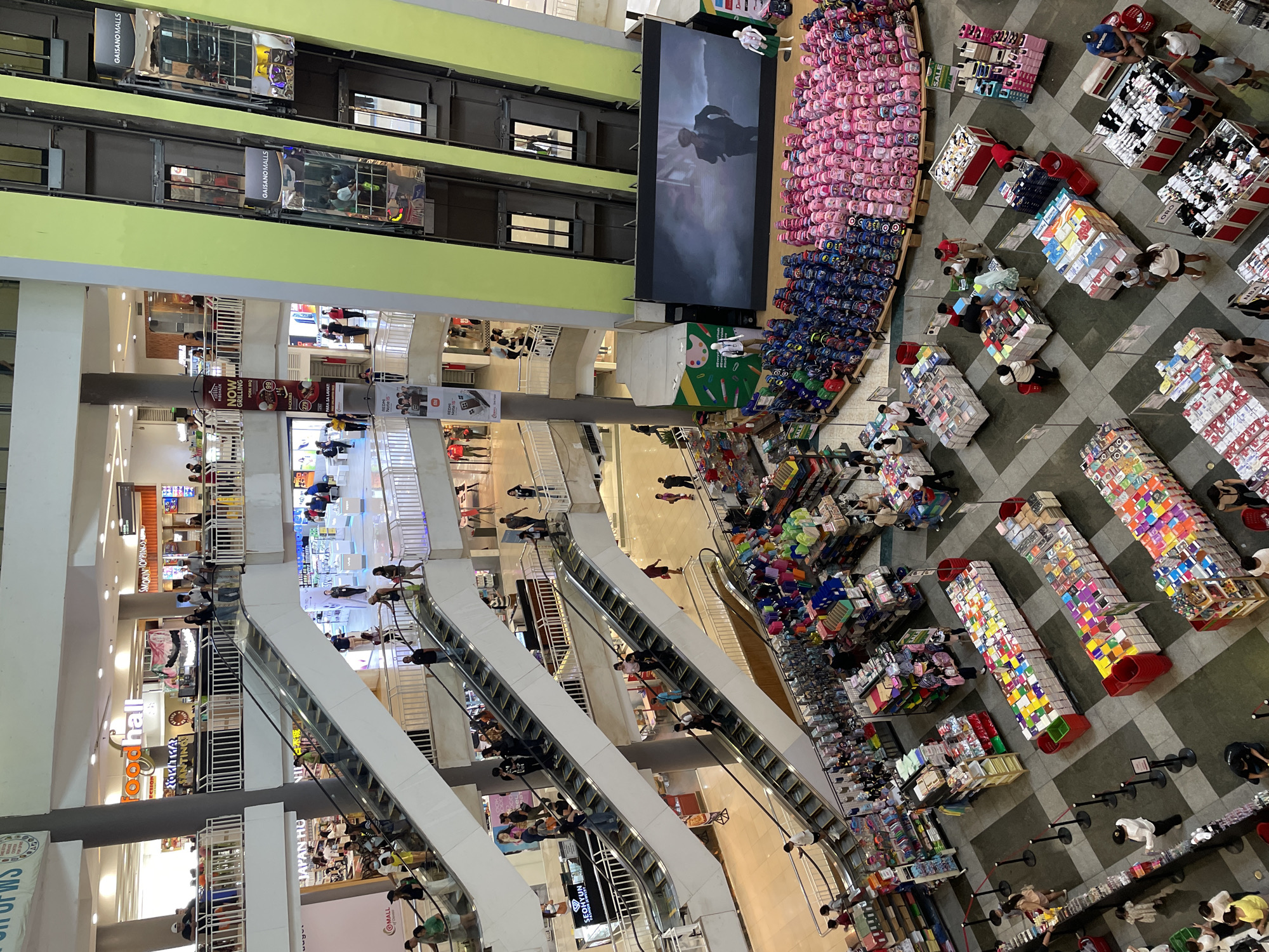 The image depicts the interior of Gaisano Mall in Davao City, Philippines, captured from an elevated perspective, likely from one of the upper floors looking down into the central atrium. The scene is a bustling commercial environment with multiple levels interconnected by escalators.
In the foreground, several rows of merchandise are displayed on the ground floor. These include stacks of colorful boxes, possibly containing stationery or school supplies, and a large arrangement of backpacks of various colors and designs, predominantly blue and pink, spread out on the floor. A large screen displays a cinematic scene, and mannequins are placed near the backpack display.
Moving upwards, multiple escalators are visible, with people ascending and descending. Shops and other commercial establishments are discernible on different levels, with signage indicating various brands and services. Some of these include "food hall" and "NOW GRILLING PORK BBQ." Further up, more shops and walkways are visible.
Elevators with glass fronts are also present, showcasing people inside as they move between floors. The architecture of the mall features a modern design with clean lines, metal railings, and a predominantly light-colored interior. The overall atmosphere suggests a typical busy shopping day in a large mall.