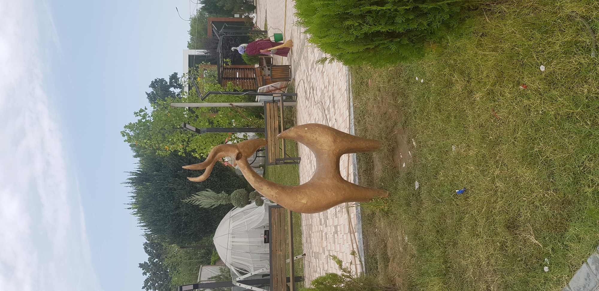 A golden statue of a stylized gazelle stands in a grassy area, its head tilted up with curved horns. Behind the statue, a paved walkway with a herringbone pattern is visible, lined with wooden benches. To the left, a white tent structure and a ladder are present. In the background, there are trees and a covered seating area. A woman wearing a headscarf and a patterned dress is sweeping the walkway with a broom and carrying a green bucket. The environment appears to be a park or outdoor public space with a mix of natural elements and man-made structures. The sky is partly cloudy.