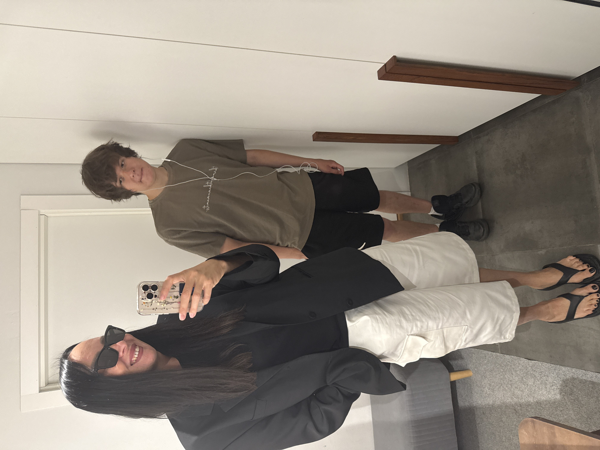 The image depicts two individuals in what appears to be a fitting room or dressing area. On the left, a person with long dark hair and sunglasses is taking a selfie with a smartphone. They are wearing a black blazer over a black t-shirt and loose white shorts, paired with black flip-flops. To the right, a young person with brown hair is standing, leaning against a white wall, wearing a tan long-sleeved shirt and black shorts, with white earbuds in. The setting features a grey bench or sofa on the left and a dark tiled floor. There are no visible indicators to suggest the location is in Chaikovsky, Russia.