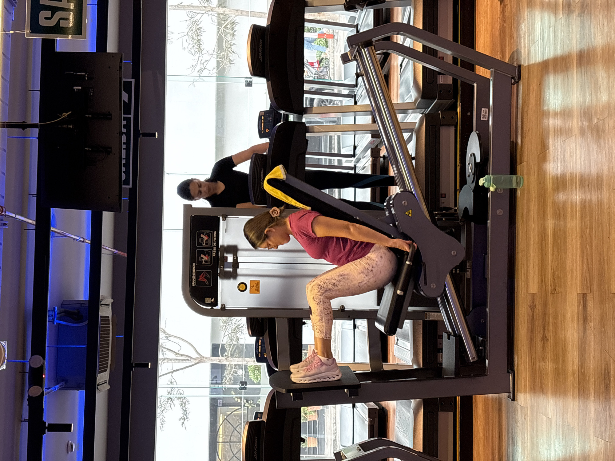 In San Isidro, Peru, at a gym, a woman is performing a leg press exercise. She is seated on a leg press machine, her feet positioned on the platform, and her legs are extended. She is wearing a pink t-shirt and patterned leggings. To her left, another woman in a black t-shirt stands near a treadmill, observing. The gym features several treadmills in the background, along with large windows offering a view of the city. A water bottle is visible on the floor near the leg press machine. The environment is well-lit with artificial lighting.