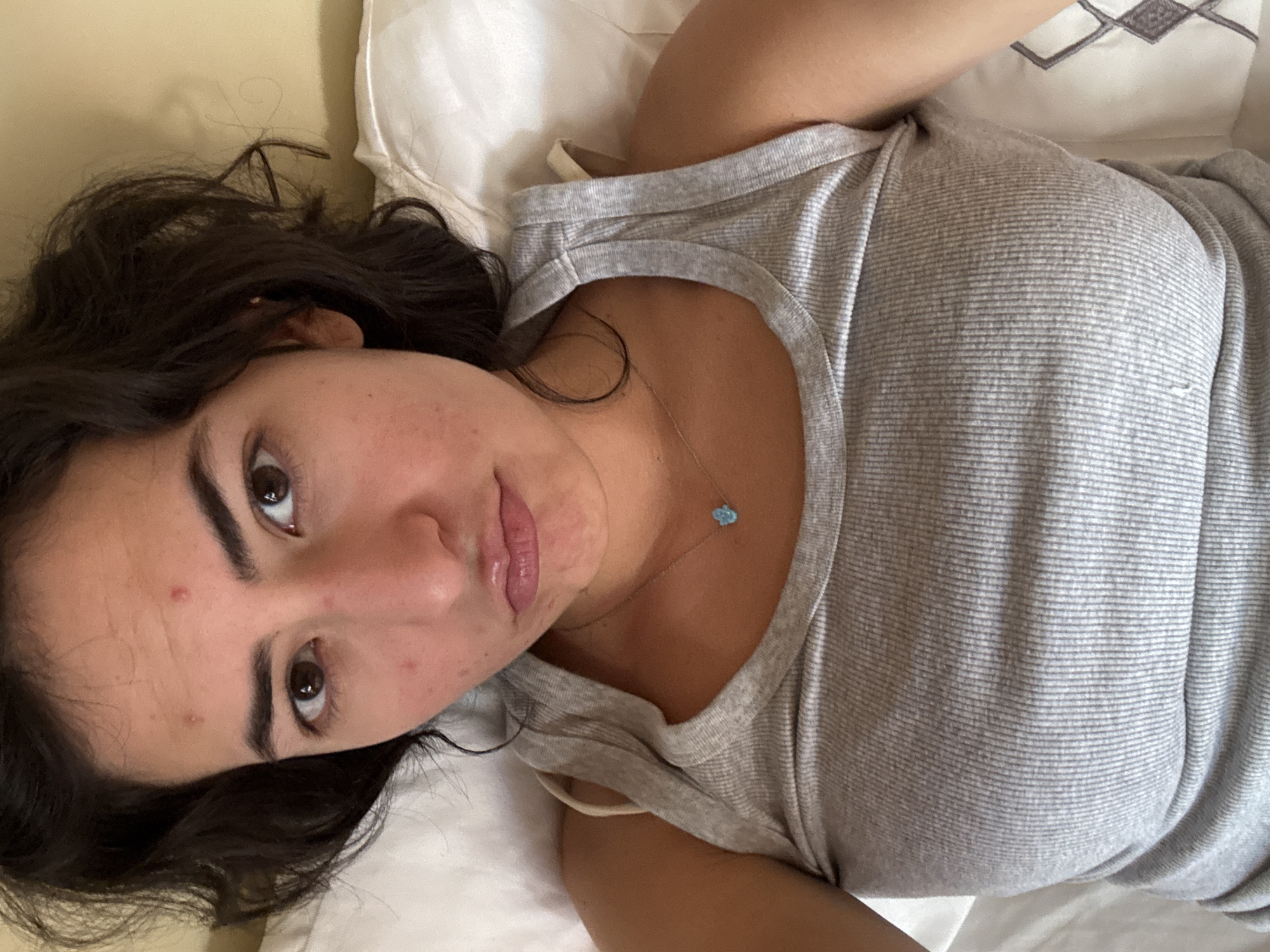 A woman with dark hair and brown eyes lies on a white pillow. She is wearing a gray tank top and a necklace with a blue pendant. Her skin appears to have some blemishes. The background is out of focus, but appears to be bedding or a pillow. The photograph is taken from a top-down perspective. There is no information to determine that the photo was taken in the city of Puebla, Mexico.