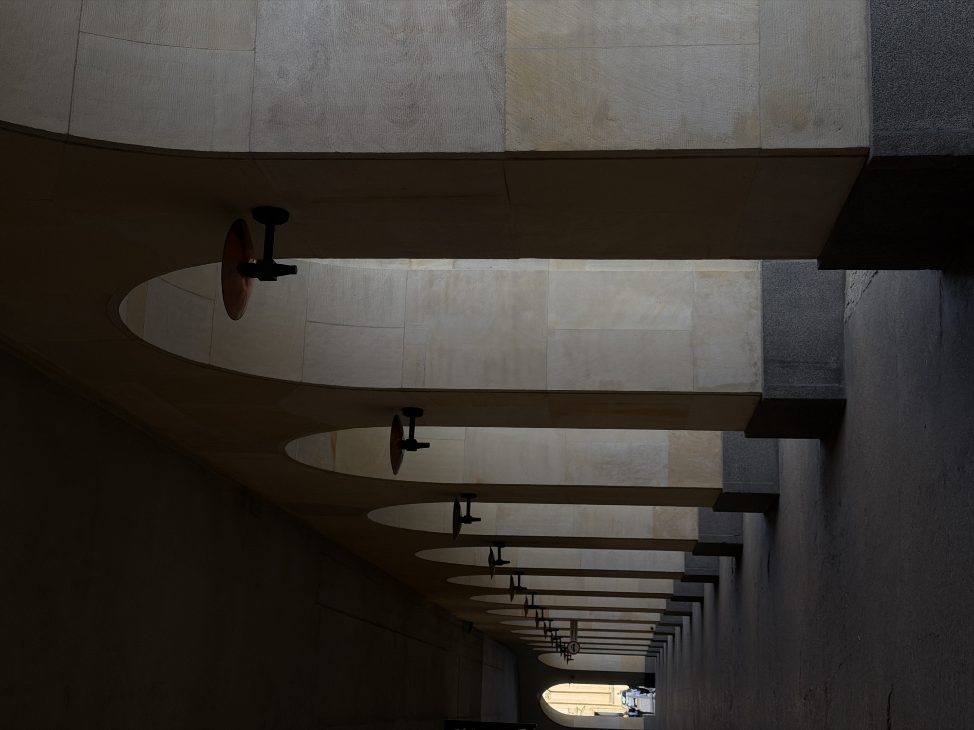 A low-angle shot depicts an arcade with a series of arches. The ceiling is constructed from light-colored stone or concrete. Attached to the ceiling within each arch are dark-colored, disc-shaped objects mounted on short, black posts. A dark grey wall extends along the right side of the arcade. The arcade opens to an area with natural light at the far end, where glimpses of vehicles and structures are visible.