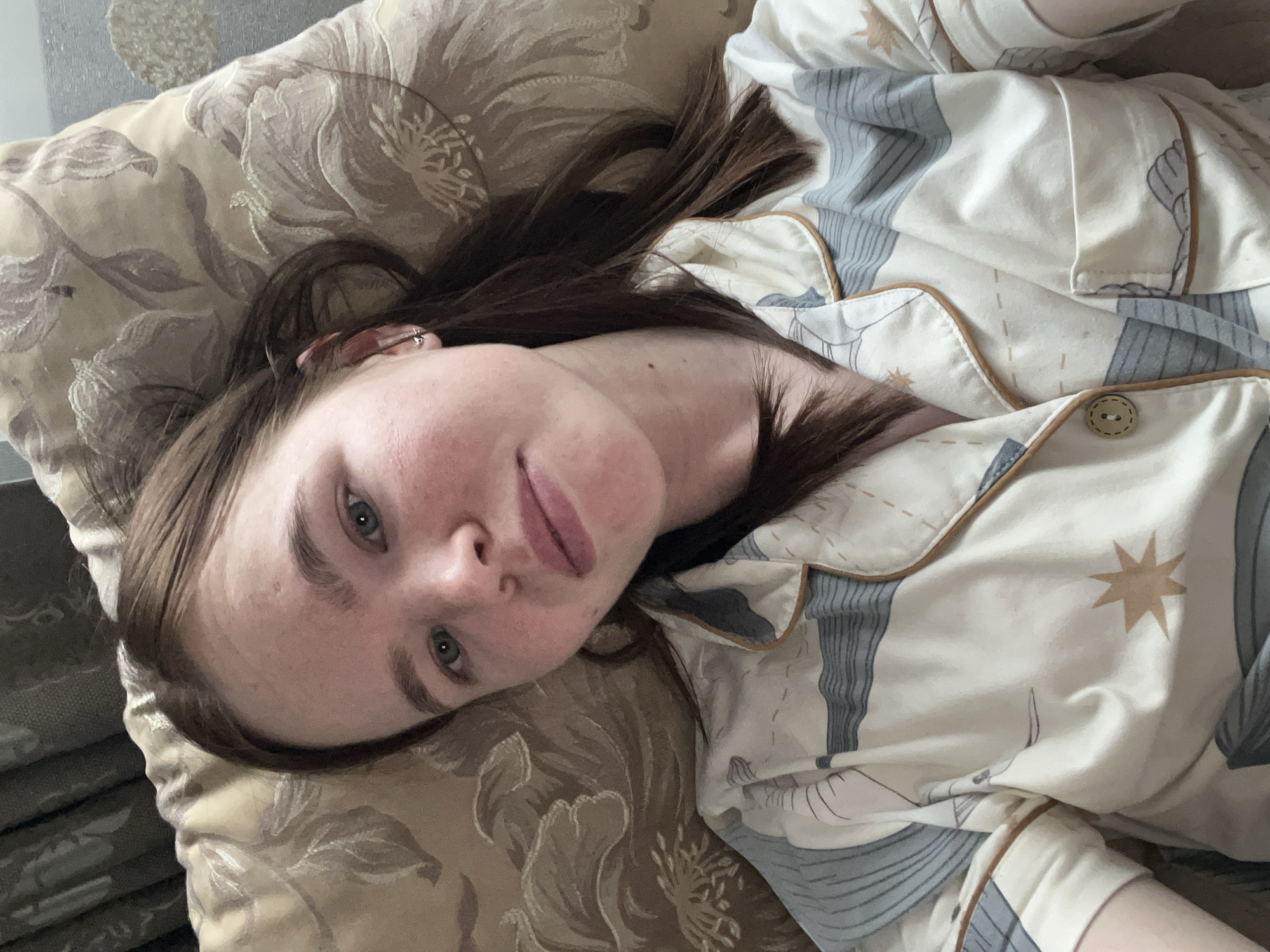 A young woman with brown hair and fair skin lies on a pillow with a floral pattern, wearing patterned pajamas with a button. The location cannot be determined as Chaikovsky, Russia.