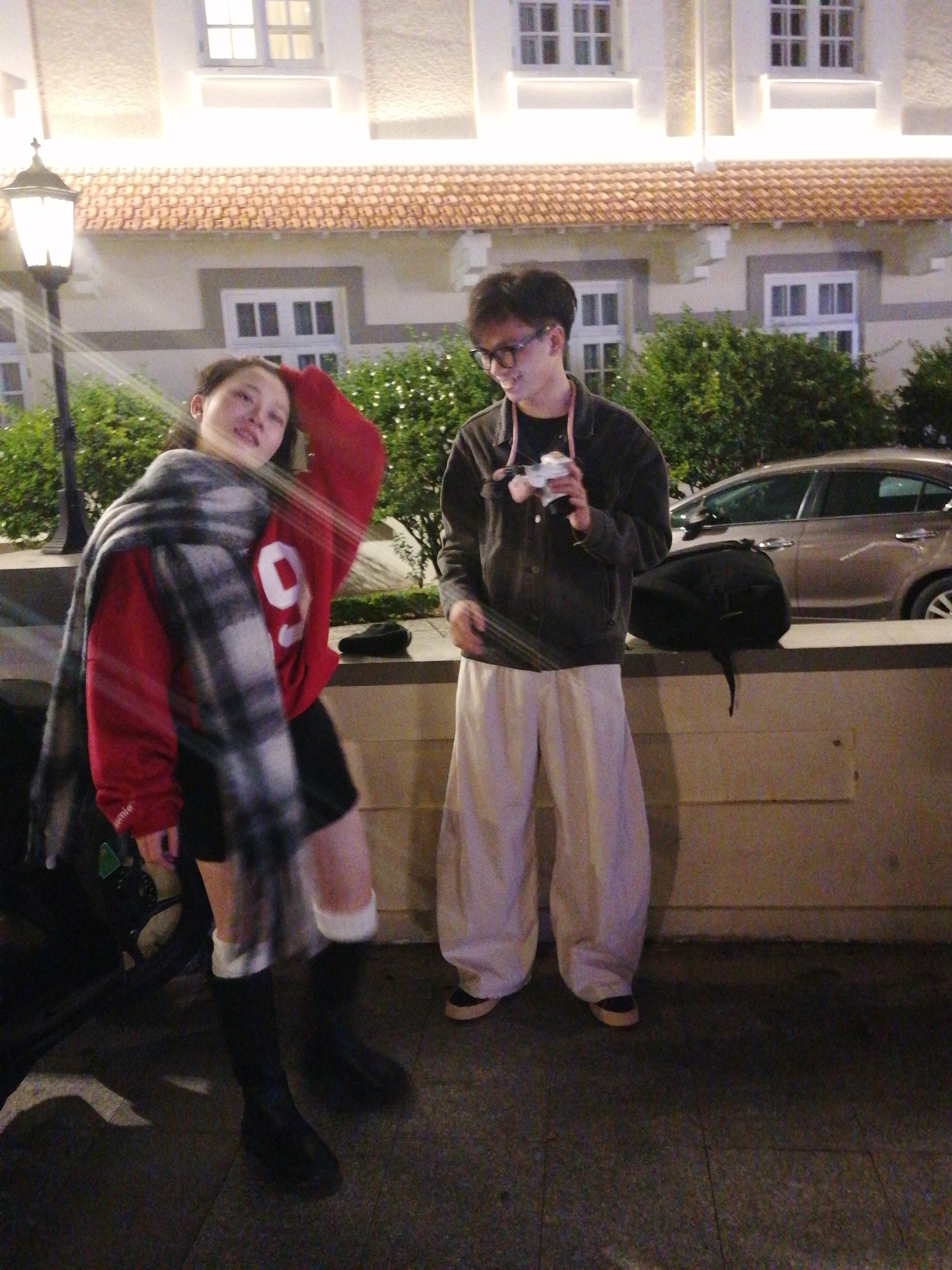 Two individuals, a female and a male, are posing for a photograph in the city of Da Lat, Vietnam. The female, positioned on the left, wears a red sweatshirt with a white number "9" on it, black shorts, knee-high black boots, and a large checkered scarf. She has her left arm raised over her head and appears to be mid-motion, with her body angled to the side. The male, on the right, is dressed in a dark jacket, a black t-shirt, and loose-fitting light-colored pants. He is holding a camera in his right hand and appears to be looking towards the camera. They are standing outdoors in front of a building with architectural details suggestive of colonial influences, characterized by a terracotta tiled roof and white window frames. Green bushes are visible behind the individuals, and a parked car can be seen in the background. A lamppost is also present on the left side of the frame. The lighting suggests it is nighttime or late evening.
