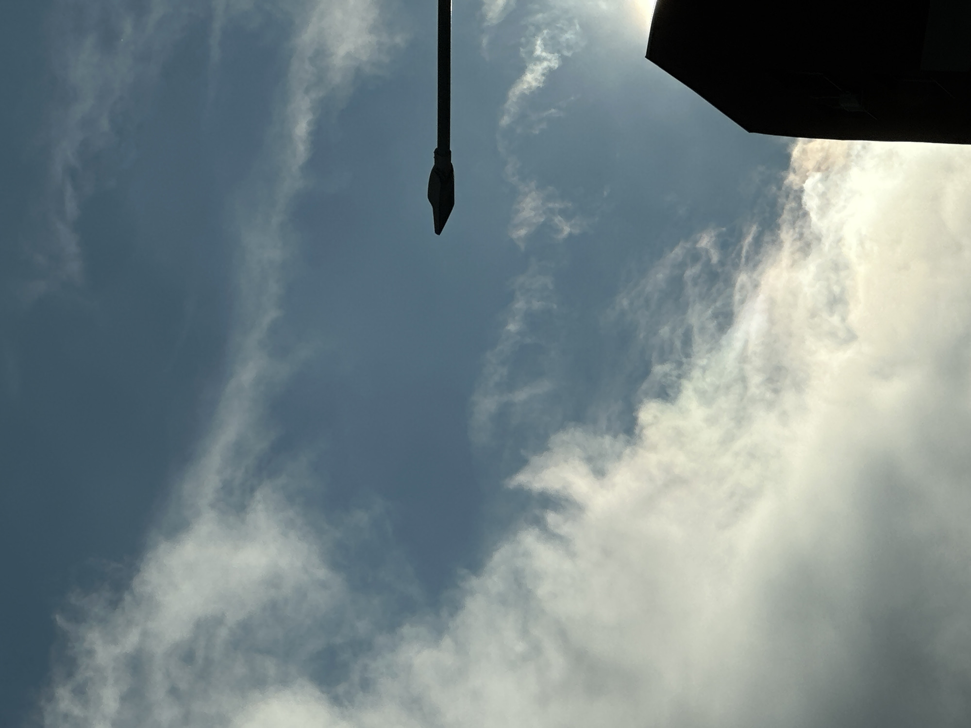 The image shows a view looking up at the sky. The sky is partially cloudy, with a mix of light blue and white clouds. A black, pole-like object hangs down from the top center of the frame, and a dark, angular object is visible in the top right corner, possibly the edge of a building. The location is likely Sofia, Bulgaria, given the context, although no specific landmarks are visible to confirm this. There are no people visible in the frame.