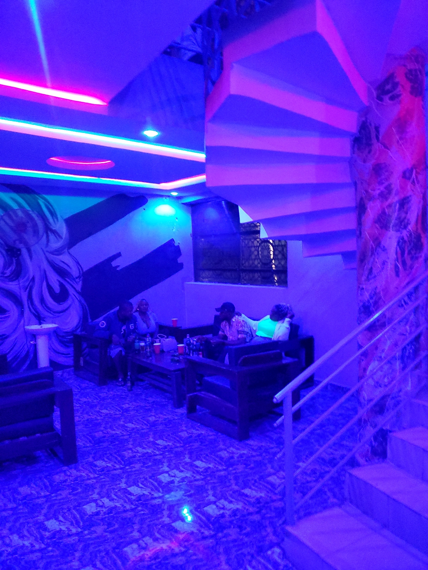 The image captures an indoor scene, likely a lounge or nightclub, illuminated by vibrant blue and pink neon lighting. In the foreground, a spiral staircase with white railings ascends along a marble-patterned wall. The floor is carpeted with a similar marble pattern. To the left, a man and a woman are seated on a dark wooden bench. A table in front of them holds several red plastic cups and bottles of drinks. Further back, another man and woman are seated on a dark sofa, with the man leaning on his hand. The walls feature a large abstract mural with black and white elements. A white pedestal sink is visible to the far left. The overall ambiance suggests a modern, dimly lit entertainment venue. The city of Jos, Nigeria, is mentioned as the location for this scene.