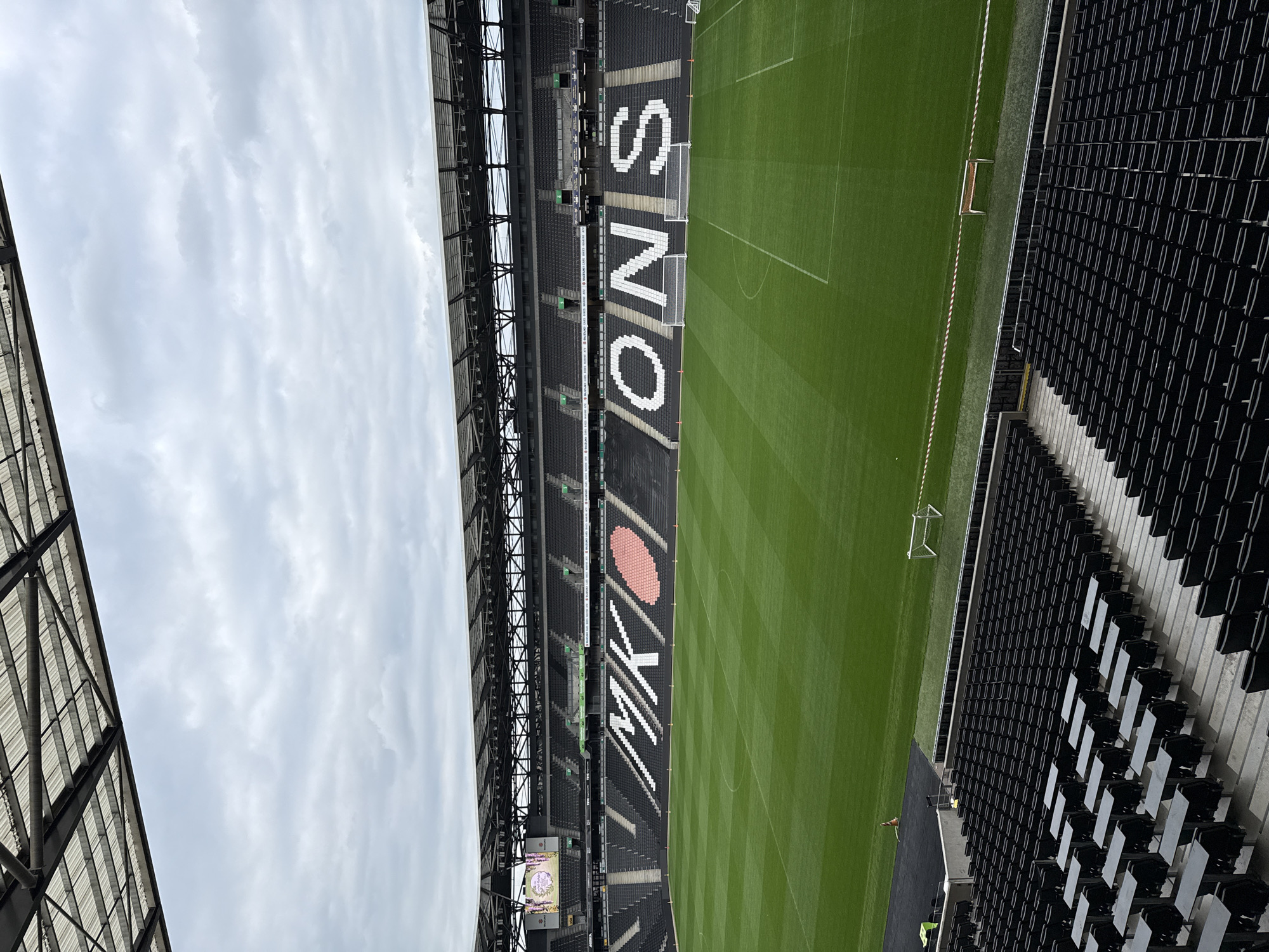 An outdoor photograph depicts a partial view of a stadium in Milton Keynes, United Kingdom, presumably Stadium MK, with a portion of the football pitch and the stands visible under an overcast sky. The grass of the football pitch is meticulously manicured with visible mowing patterns. Two goalposts stand at either end of the visible section of the pitch. The seats in the stands are predominantly black, with white block lettering spelling out "MK DONS" running vertically down the seating. A large, heart-shaped graphic is integrated into the "MK" part of the lettering. To the left, a scoreboard or large screen is visible, though the image displayed on it is indistinct. The stadium's architecture includes a complex network of metal supports and roofing. There are few people visible in the lower right corner of the image, and their actions are not clear.
