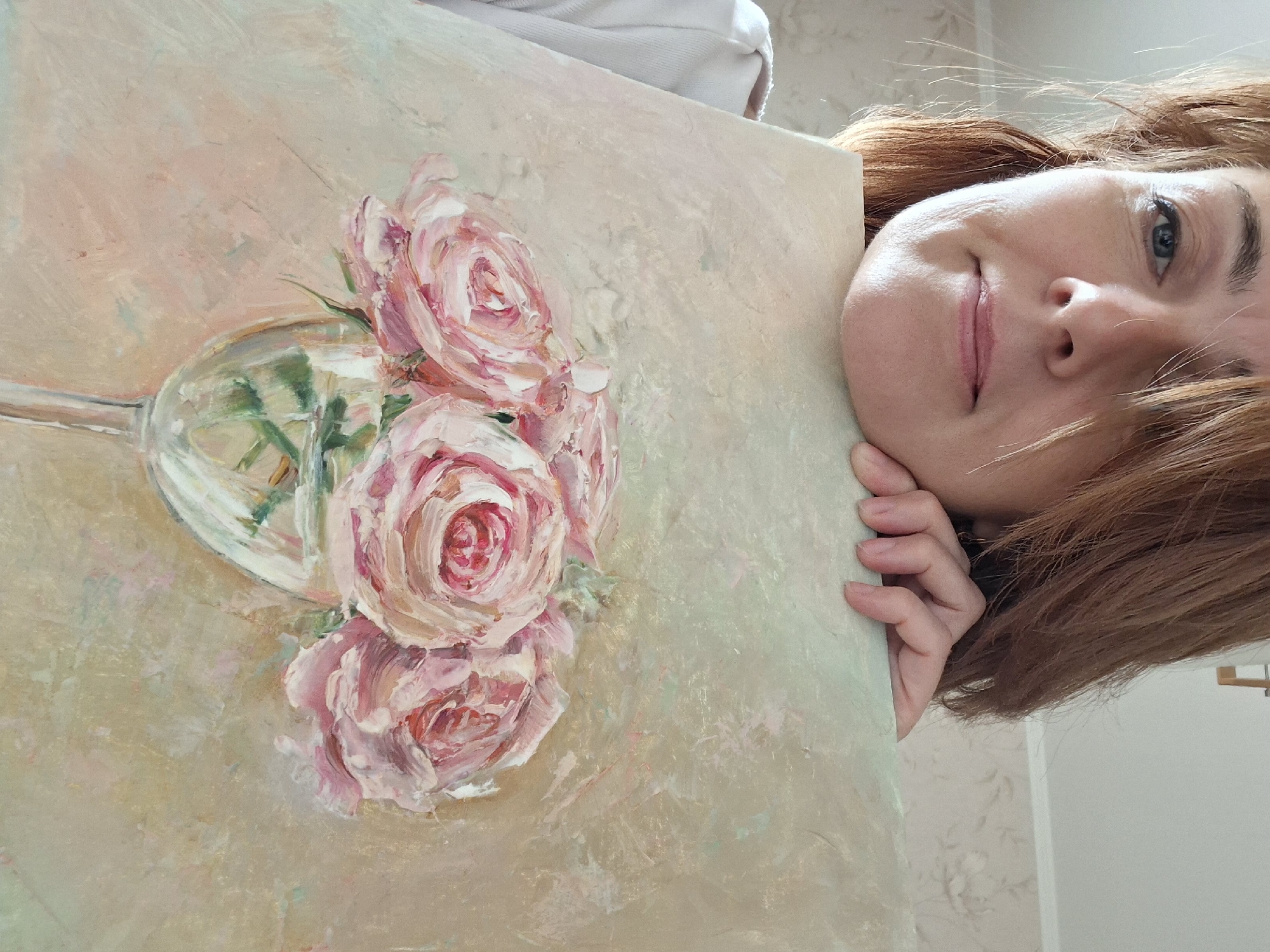 A woman with auburn hair is holding a painting of pink roses in a glass vase. She is looking at the camera with a slight smile. The painting appears to be an oil painting with thick brushstrokes. The background of the painting is a light beige color with hints of pink and green. The woman is wearing a white shirt. The image is taken indoors, with a patterned wallpaper visible in the background. The lighting suggests it is daytime. The woman's face is partially obscured by her hair and her hand resting on the painting.
