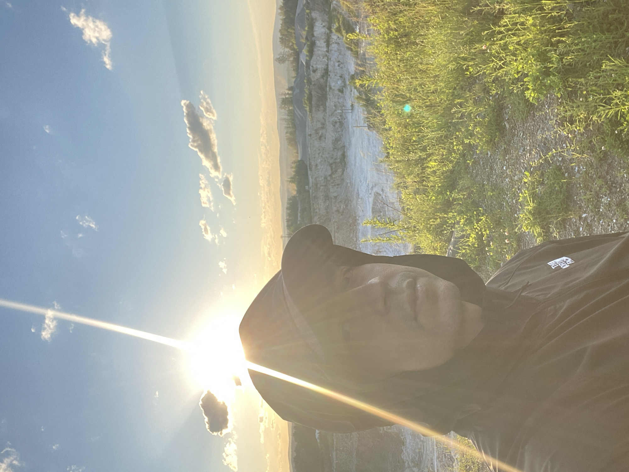 A selfie taken by a person wearing a black hooded jacket and a black baseball cap is featured in the foreground. The person's face is partially obscured by the cap and a strong sunbeam. Behind them, a quarry is visible, with layered rock formations and sparse vegetation. The sky is clear blue with a few scattered clouds, and the sun is setting, casting a warm, golden light over the landscape. The overall scene suggests an outdoor setting, possibly in a rural or industrial area. The specific location in Yekaterinburg, Russia, is not evident from the image itself.