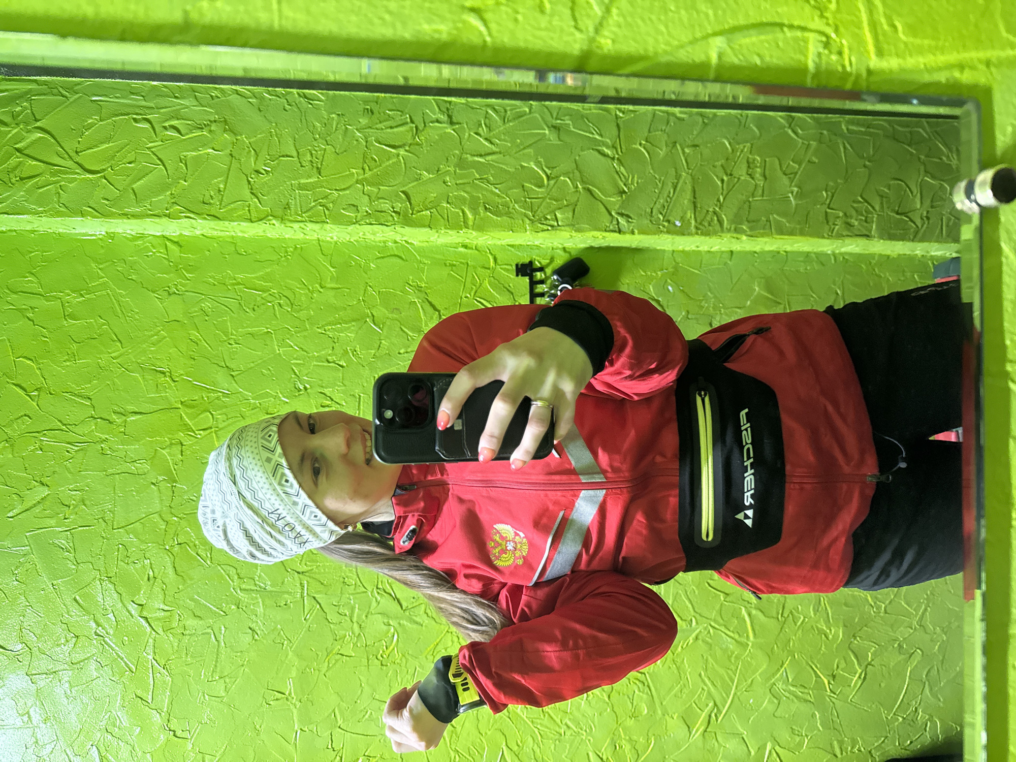A woman is taking a selfie in front of a mirror, with a bright green textured wall behind her. The wall appears to be in a room with fluorescent lighting. The woman is wearing a patterned white headscarf, a red jacket with reflective silver stripes and the emblem of Russia on the left chest, and a black Fischer fanny pack. She is holding a black smartphone in her right hand. Her left hand is visible with a black and yellow smartwatch. The mirror reflects the room, revealing a metallic fixture on the right edge of the mirror. The room's wall is very textured. While the location is not discernible from the image, it is stated in the prompt to be the city of Chaikovsky, Russia.
