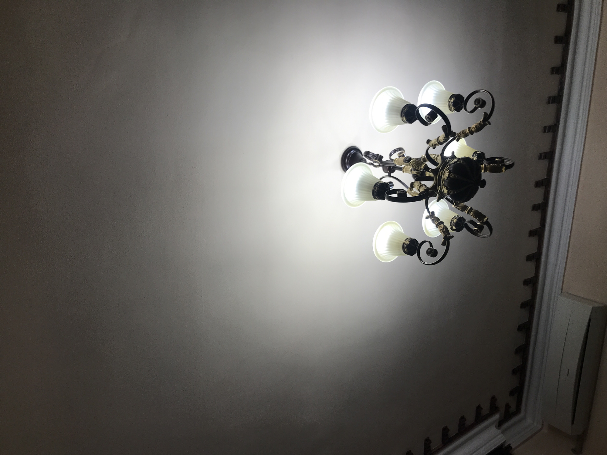 A low-angle shot of a ceiling in Yerevan, Armenia, featuring an ornate chandelier with multiple white lamps. The ceiling appears to be a light gray color, possibly painted plaster or drywall, with some uneven lighting. The chandelier is positioned slightly off-center to the right, casting a brighter area around it. Near the right edge of the frame, part of the room's architectural detailing is visible, including a section of wall trim or molding and a white air conditioning unit or wall heater.
