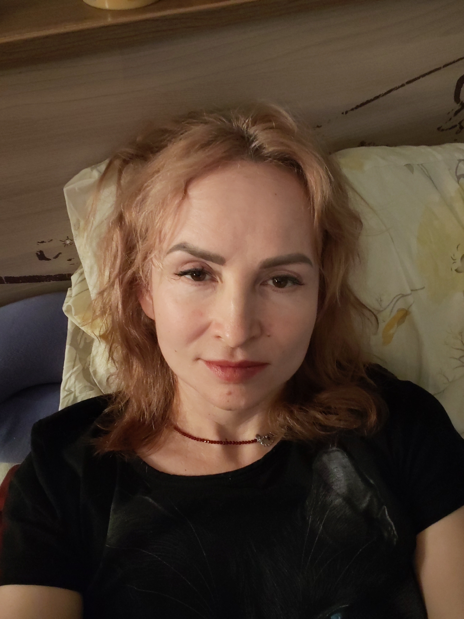 A fair-skinned woman with strawberry blonde hair lies in a bed with a patterned pillowcase. She wears a black t-shirt with a cat design and a red beaded necklace. The location of the image is not evident from the photograph.