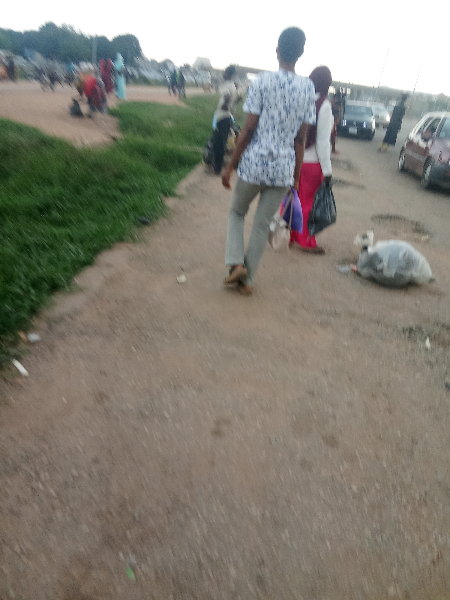 The image captures a street scene in Kagini, Nigeria. In the foreground, a person is walking away from the camera, wearing a patterned shirt and light-colored pants. The person is carrying two bags, one purple and the other black. To the right of this individual, another person is walking, wearing a white top and a pink skirt. Further down the road, several vehicles are visible, including a dark-colored sedan and a red station wagon. The ground is a dirt road with patches of green grass along the left side. In the background, more people and structures are indistinctly visible, suggesting a populated area. There is also a large, grey, trash bag lying on the ground to the right of the person in the patterned shirt. The overall lighting suggests it is late afternoon or early evening.
