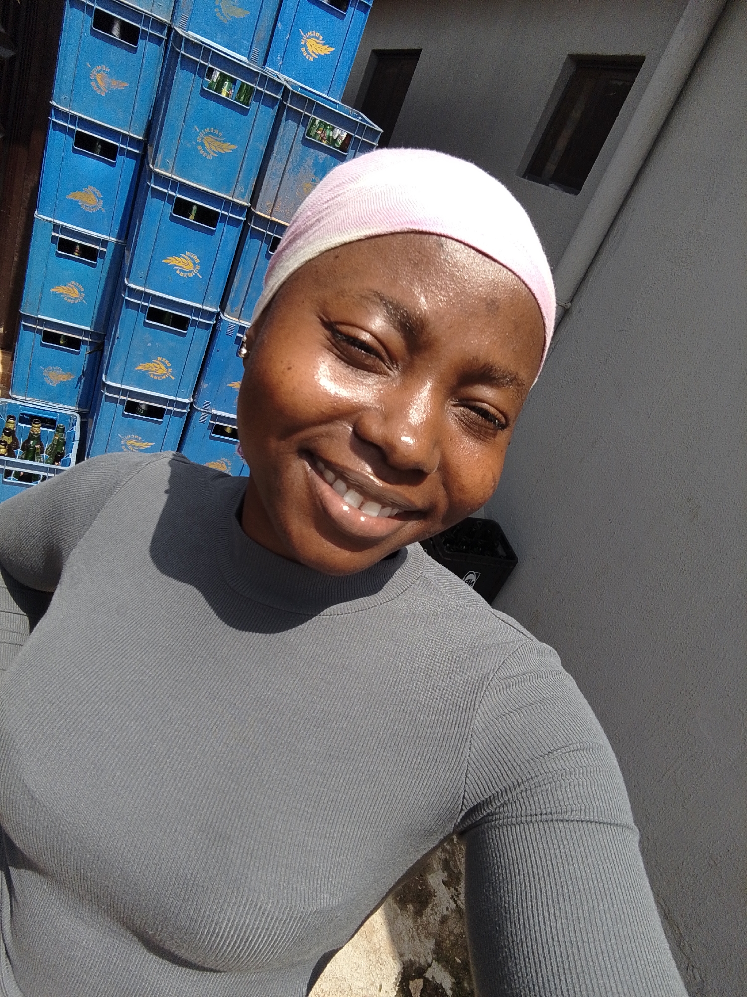 A young woman with dark skin and a pink headscarf smiles at the camera. She wears a grey, ribbed long-sleeved shirt. Her face is shiny, suggesting sweat or oil, and she is outdoors in what appears to be a sunny environment. Behind her, several stacks of blue plastic crates, possibly for beverages, are visible. Some of these crates contain bottles. A grey, textured wall with two dark, rectangular window openings is to her right. The scene suggests a bright, warm day. The overall emotion conveyed is happiness and a sense of well-being.