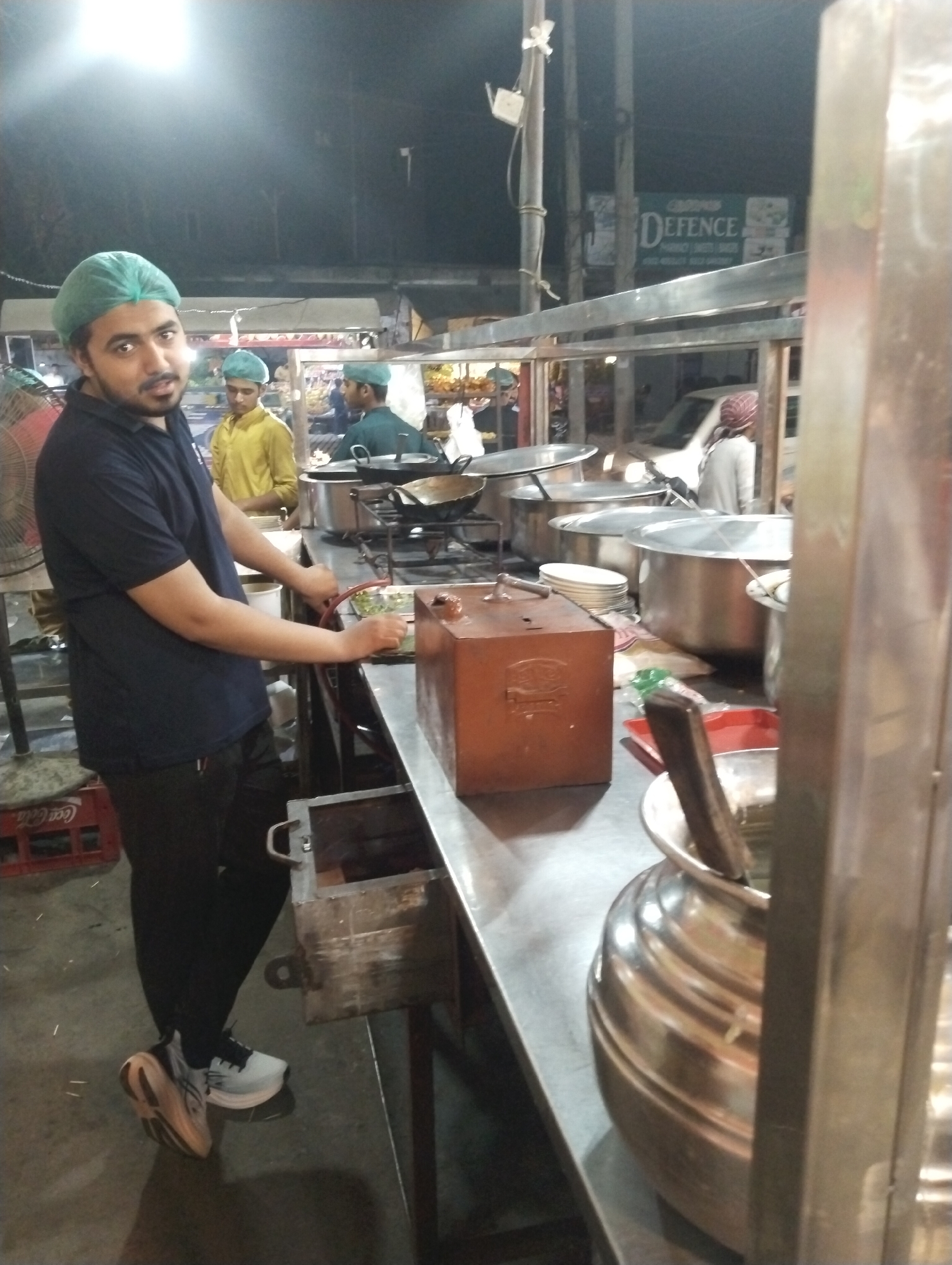 The image captures a nighttime scene at an outdoor or semi-outdoor food stall or small eatery, likely in Heir, Pakistan. The primary subject is a young man in the foreground, wearing a dark blue polo shirt, black pants, and a light green hairnet/cap. He has a beard and is looking directly towards the camera with a neutral expression. He stands beside a stainless steel counter, where his hands are near a red hose-like object and a red plastic tray containing green ingredients, suggesting he is involved in food preparation or serving. He wears light grey and white athletic shoes with distinctive tan soles.
In the background, two other men also wear light green hairnets, indicating they are staff. One man in a yellow shirt stands behind the counter, while another in a dark green shirt is further back, seemingly engaged in cooking activities. Various large, lidded stainless steel pots, woks on burners, and stacked white plates are visible on and behind the counter, typical of a busy street food establishment. A brown wooden box, possibly for cash or supplies, sits on the counter. A standing fan is positioned to the left of the main subject, and red Coca-Cola crates are visible on the floor.
The setting is illuminated by artificial lights, with a bright overhead light source visible above the main subject. In the distance, a white car is parked, and utility poles with wires are present. A green signboard in the background clearly shows the word "DEFENCE," with other partially visible words like "PHARMACY," "SWEET," and "BAKERY," along with some phone numbers, suggesting a commercial area. The overall scene depicts a functional and bustling food preparation area during evening hours.