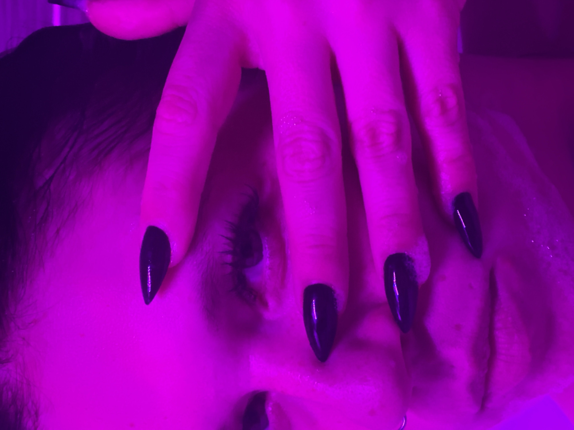 The image is a close-up, low-angle shot of a person's face, lit by intense purple light. The subject's eyes are partially obscured by their own hand, which has long, sharp, black-painted fingernails. The hand is positioned across the upper part of the face, with fingers covering the bridge of the nose and near the eyes. Some white, foamy substance, possibly soap or skincare product, is visible on the person's face and fingernails. The background is a solid, dark color with no discernible features, suggesting an indoor setting. There is no information available to locate this image in Chaikovsky, Russia.