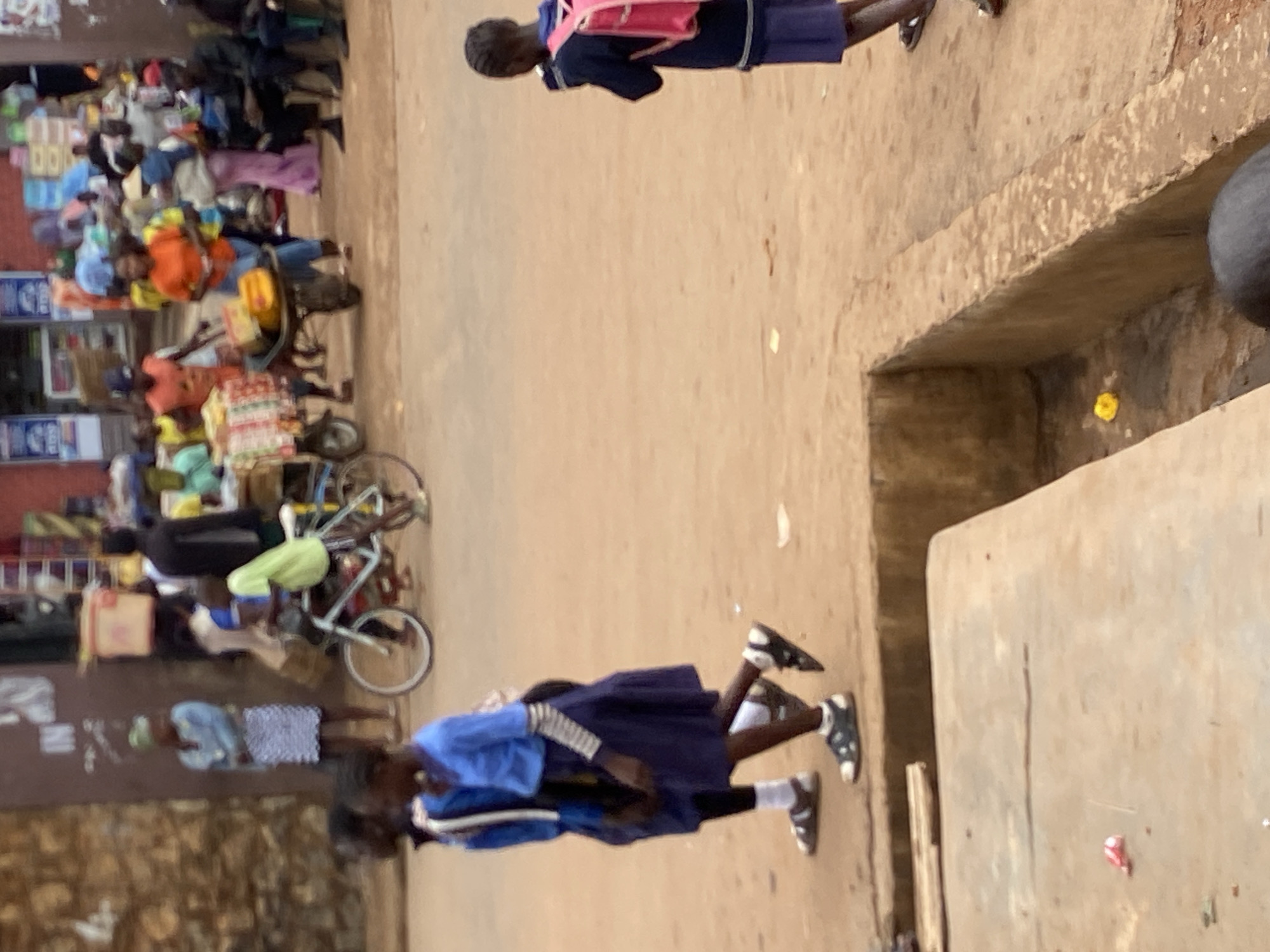 The scene is in Bukuru, Nigeria. Two schoolgirls in blue and white uniforms and black sandals are walking down a dirt street. One wears a pink backpack. On the left side of the street are people and bicycles near storefronts. Merchandise is visible in front of the stores. The street surface is dirt and brown. A concrete structure is on the right of the image.