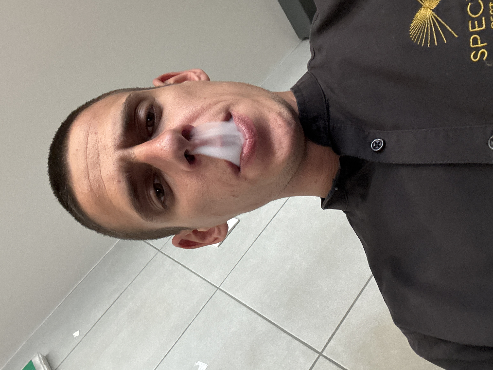 A close-up, tilted selfie captures a man exhaling smoke. The man has short, dark hair, light skin, and brown eyes. He wears a dark shirt with embroidered text/design visible on the upper right side of the frame. White smoke billows from his mouth. The background consists of light-colored tiled walls and flooring. The image does not contain any indication of Sofia, Bulgaria.