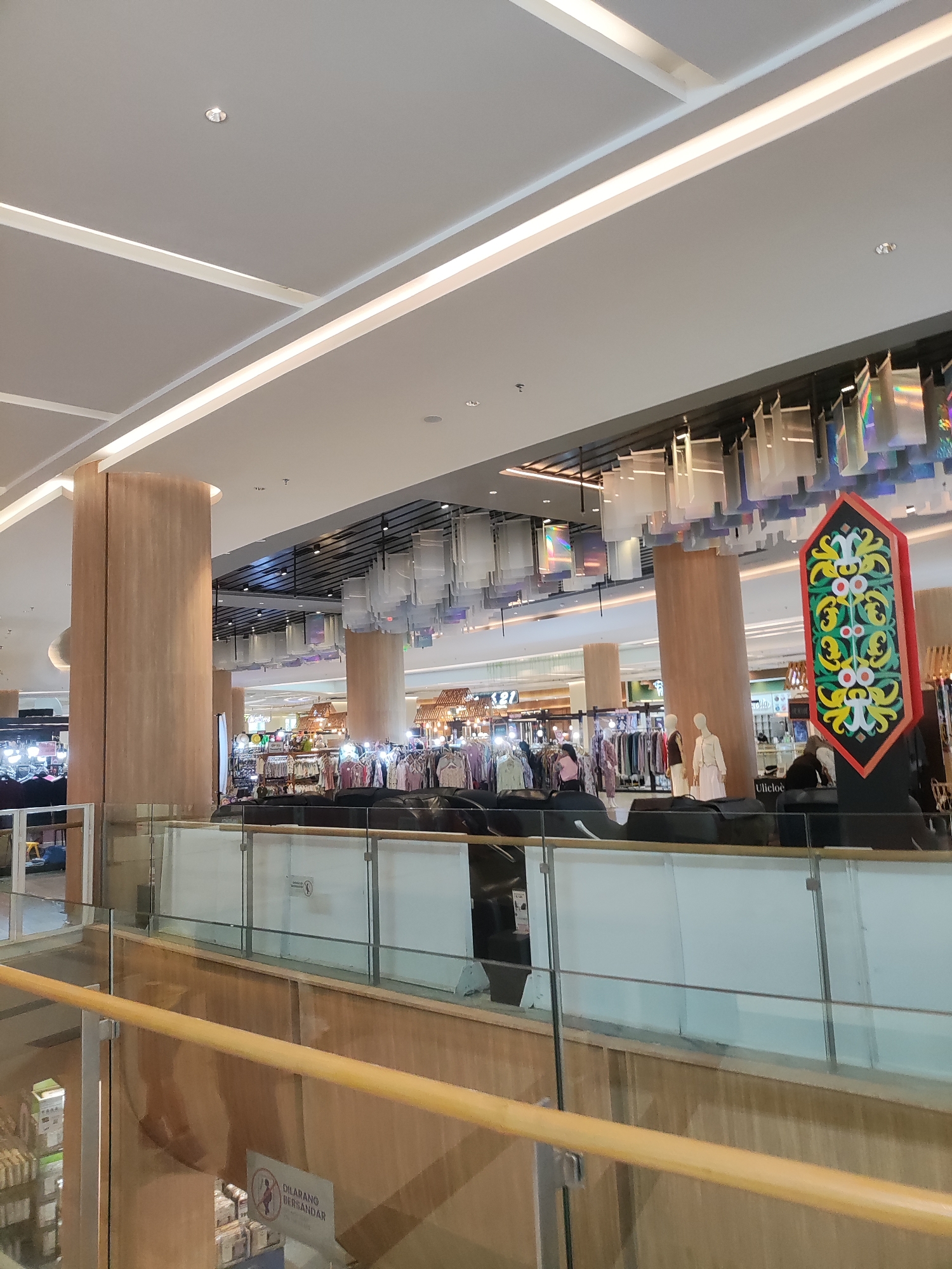 The image depicts an interior view of a shopping mall, likely in Lambangjaya, Indonesia, given the sign with Indonesian text. The scene is illuminated by bright, modern lighting integrated into the ceiling and decorative fixtures. The architecture features large, cylindrical columns clad in light brown wood-grain material and a multi-level structure with glass and wooden railings.
In the foreground, a wooden handrail runs across the lower part of the frame, leading the eye towards a lower level. On this lower level, a sign with red and green motifs, reminiscent of traditional Indonesian art, stands prominently. Further back, several stalls or shops are visible, displaying clothing on racks and mannequins. One stall has several mannequins dressed in light-colored attire. Above the shops, a ceiling installation of translucent, rectangular panels shimmers with iridescent colors, adding a dynamic visual element to the space.
In the mid-ground, a glass barrier with metal supports separates different levels of the mall. Through the glass, several rows of clothing racks with various garments are visible. A few people can be seen browsing or working in these areas, though their faces are not clear. One person is standing near a mannequin, and another is partially visible near a clothing rack. On the far right, a man wearing a dark top and light-colored trousers is seated.
The environment appears clean and well-maintained, characteristic of a modern commercial space. The overall impression is one of a bustling, well-lit shopping environment with a blend of modern design and cultural decorative elements.