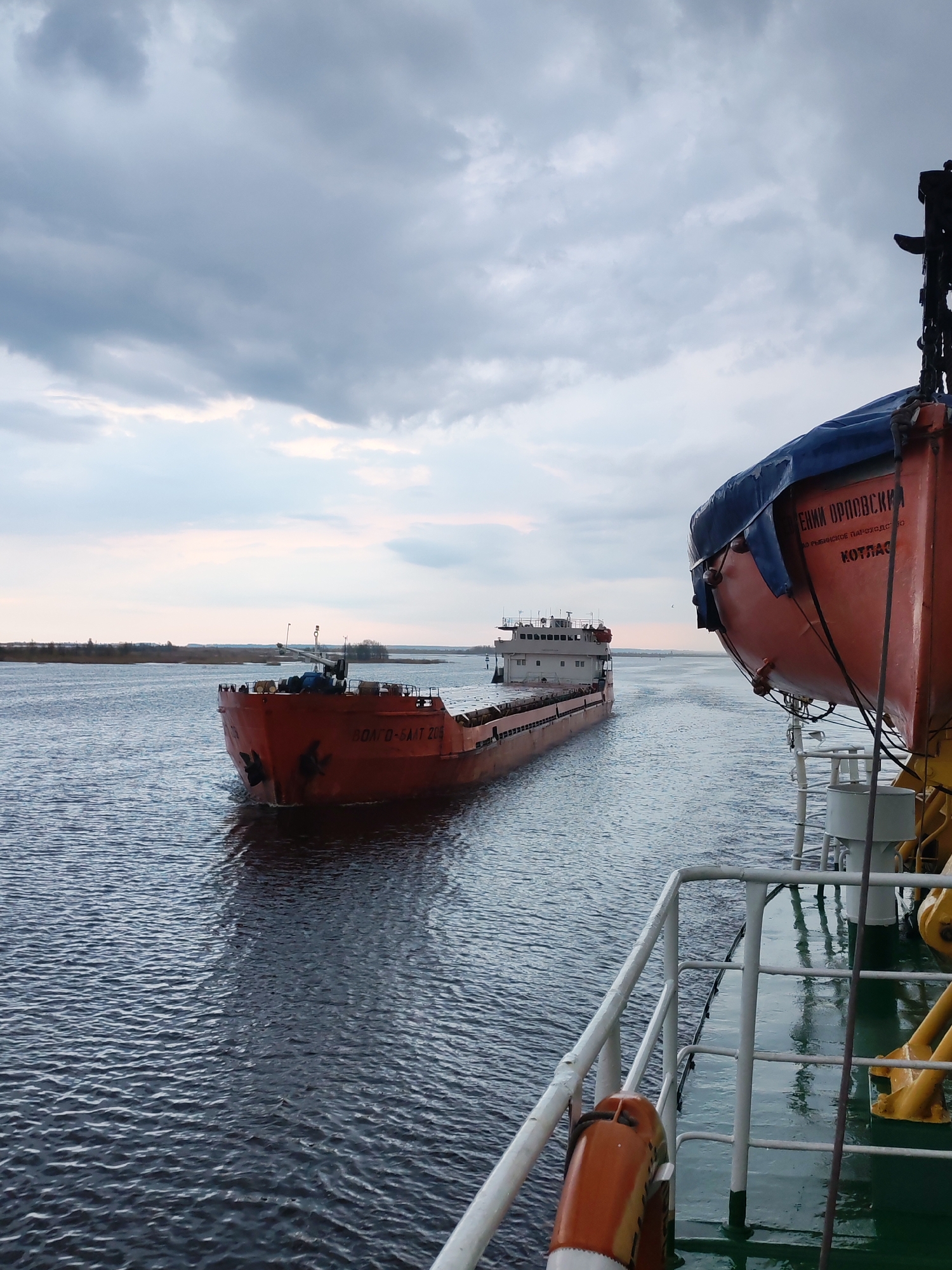 The image depicts two ships on a body of water under a cloudy sky. The ship in the foreground has a bright orange lifeboat with Cyrillic text on it. The background ship, also orange, bears the inscription "ВОЛГО-БАЛТ 208". The environment includes water, cloudy skies, and possibly a riverbank in the distance. The location is potentially near Novokemskii, Russia, based on the ship markings and context. No people are clearly visible. The primary action depicted is the movement of ships on the water.