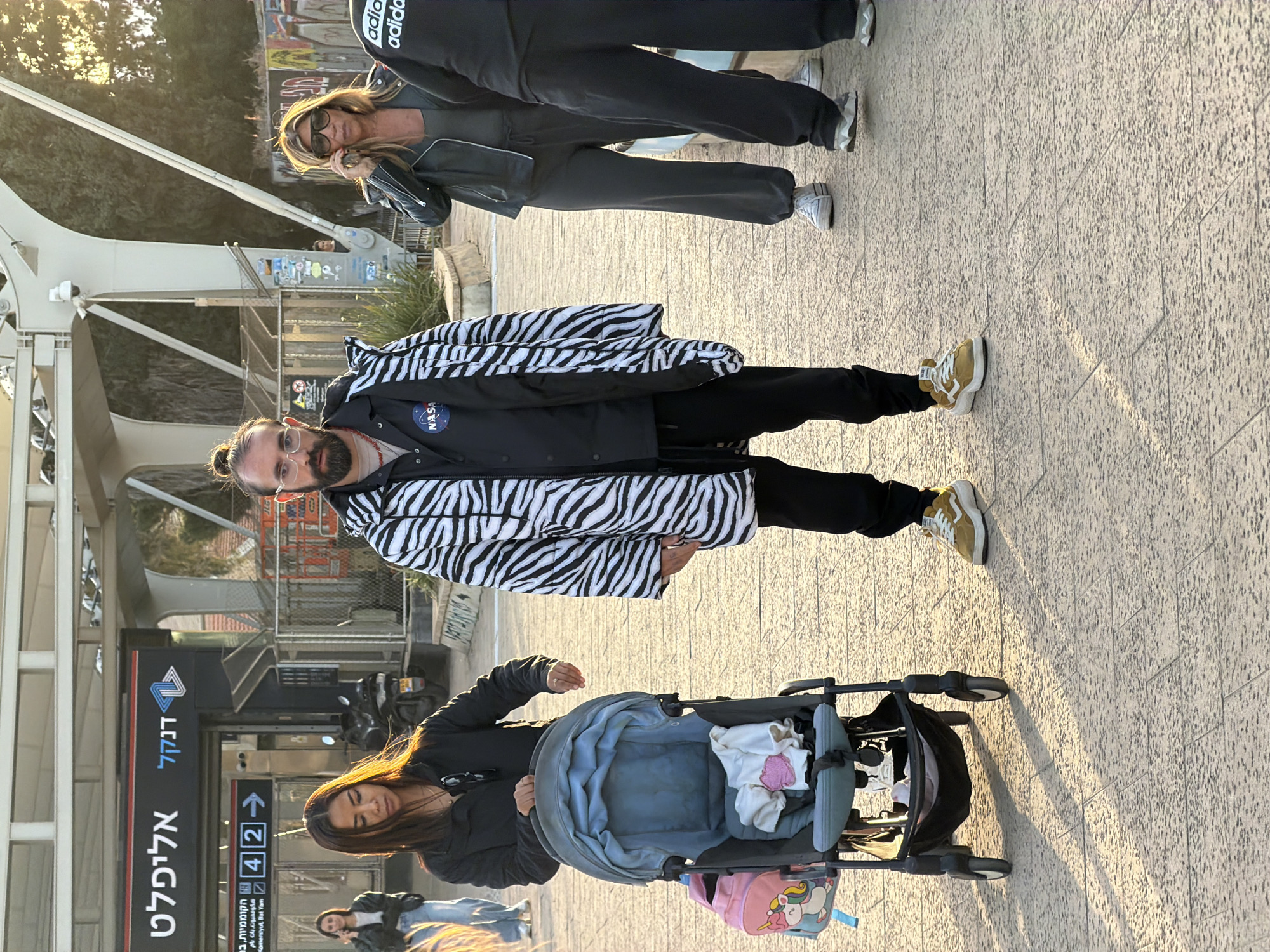 The image shows a scene in Tel Aviv, Israel, outside the Allenby station of the Red Line light rail. A man wearing a zebra-striped jacket and NASA logo is walking on a paved surface. A woman is pushing a stroller with a baby in it. Other people are walking by. The background includes a bridge structure and trees.