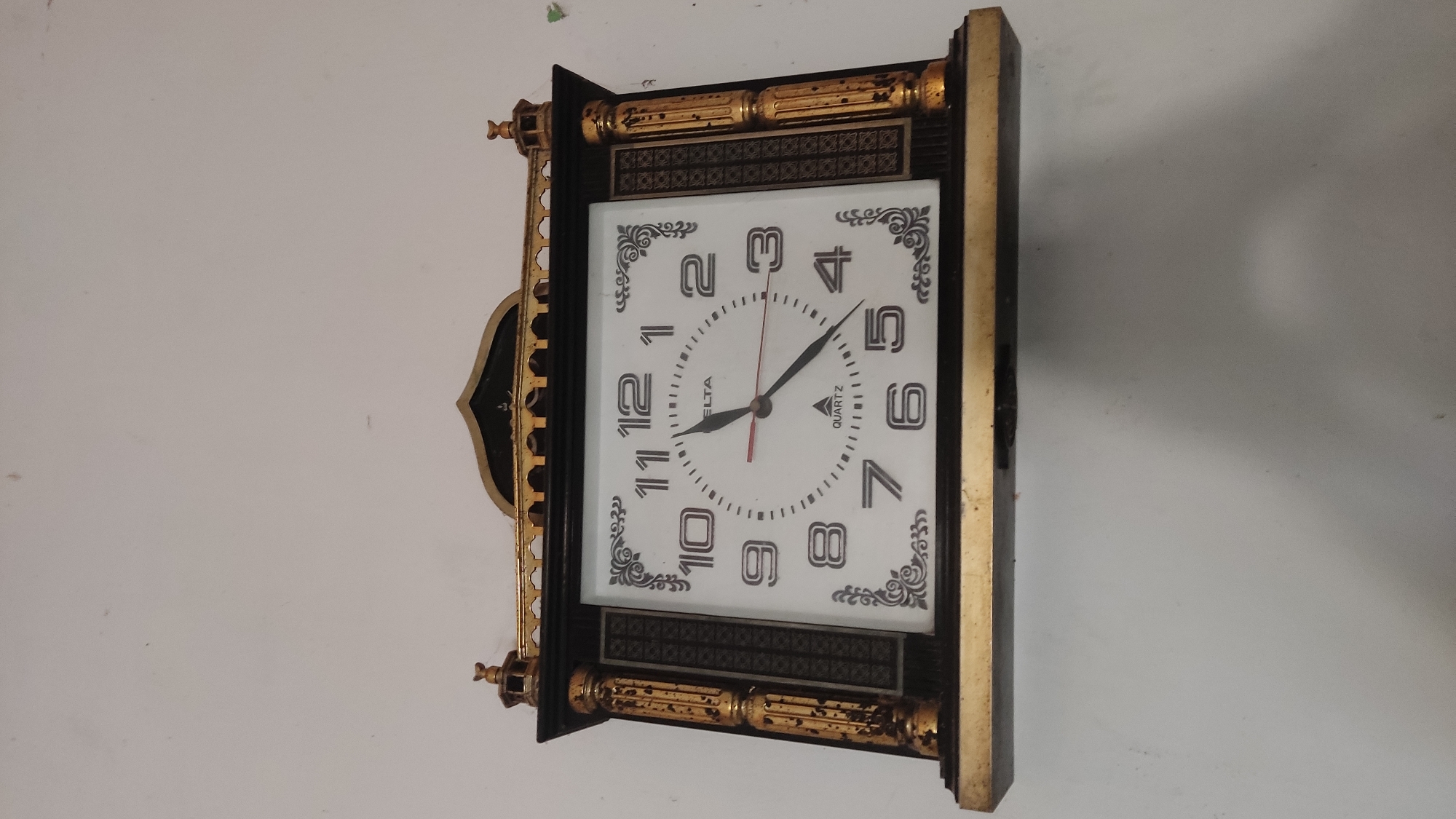 A decorative analog wall clock with a white face and black numerals is mounted on a white wall. The clock has a dark wooden frame with gold accents, including ornate columns on either side and a gold decorative element at the top that resembles an Islamic architectural motif. The clock hands indicate a time of approximately 11:25. The "DELTA" and "QUARTZ" branding is visible on the clock face. The wall appears to be unpainted or distempered. The image is taken from a slightly upward angle. The clock is likely located indoors, possibly in a home or public building. The environment suggests a typical Indonesian setting, specifically Ngadiharjo, Indonesia.