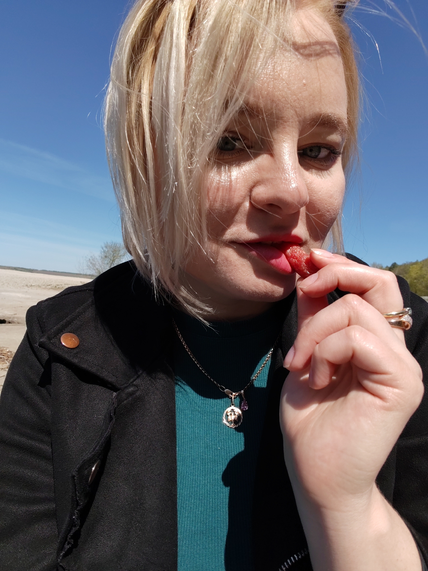 A woman with short blonde hair, wearing a black jacket over a teal turtleneck sweater and a necklace with a circular pendant and a small purple stone, is holding a red, textured fruit between her fingers and bringing it to her mouth. She has two gold rings on her ring finger. The background consists of a light-colored, sandy area that appears to be a beach or shore, a blue sky, and some distant trees. The location is Икское Устье, Russia, identified by the geographic features characteristic of the area.